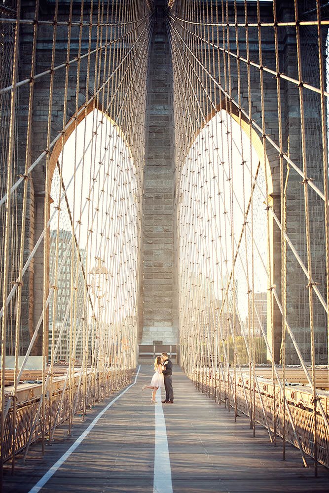 A couple standing on a Brooklyn Bridge walkway at sunset, with city buildings in the background.