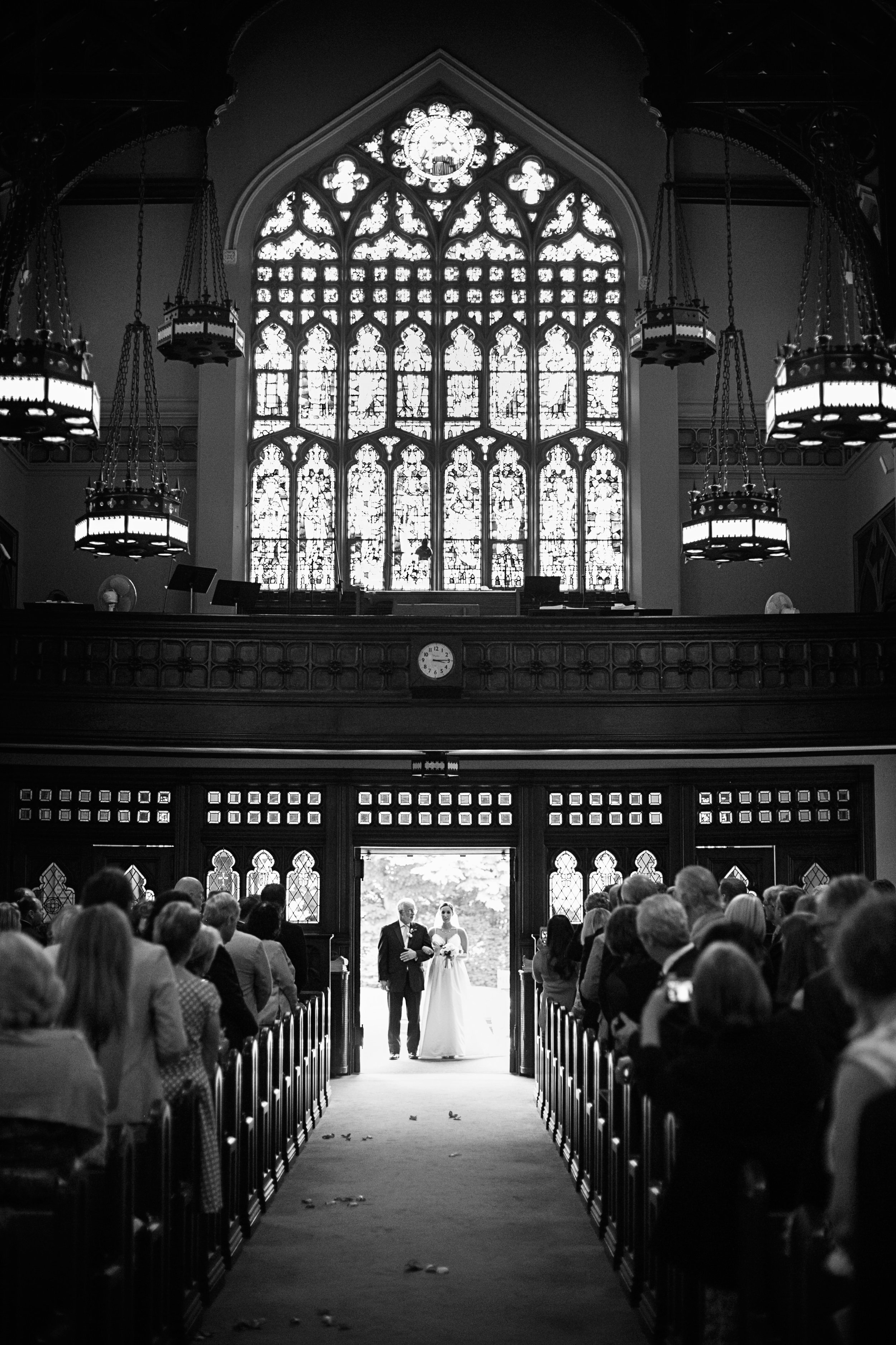 A black-and-white photo of a church interior with a large stained-glass window. A bride is walking down the aisle with an older man, possibly her father, as guests seated on both sides watch. The church has gothic-style architecture with chandeliers 