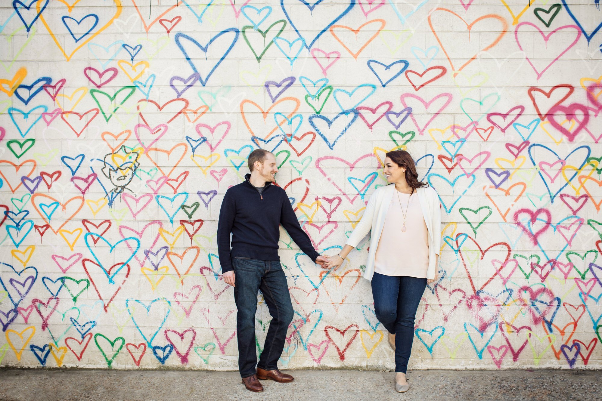 A man and woman holding hands and smiling at each other in front of a colorful wall covered in variously colored heart and face graffiti.
