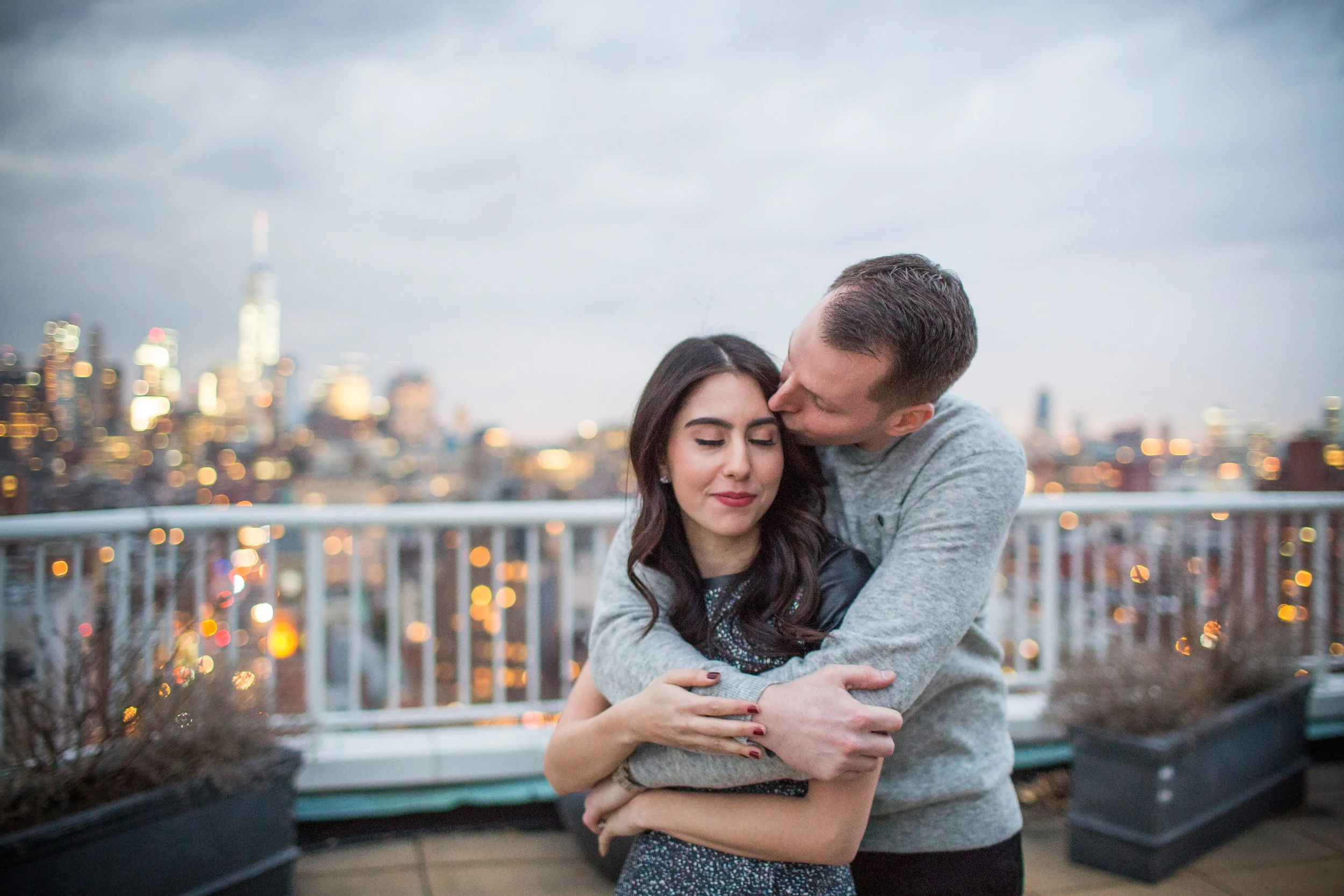 A couple hugging on a rooftop with a city skyline in the background, late evening.