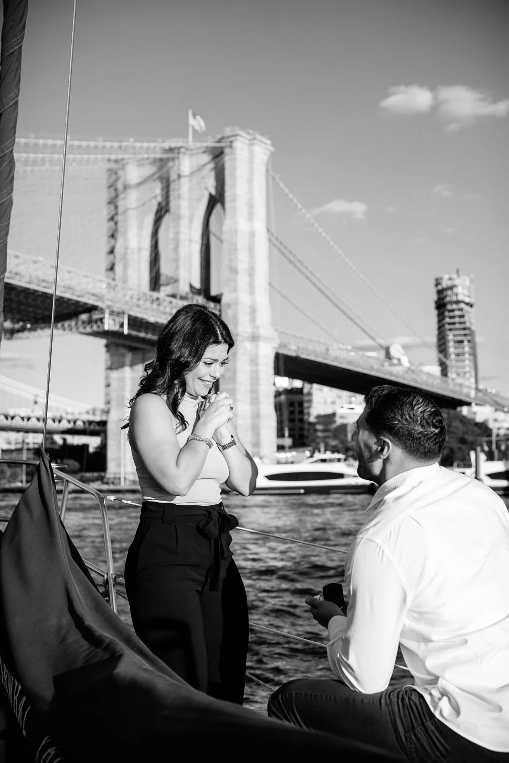A man on one knee proposes to a woman on a boat with the Brooklyn Bridge in the background.