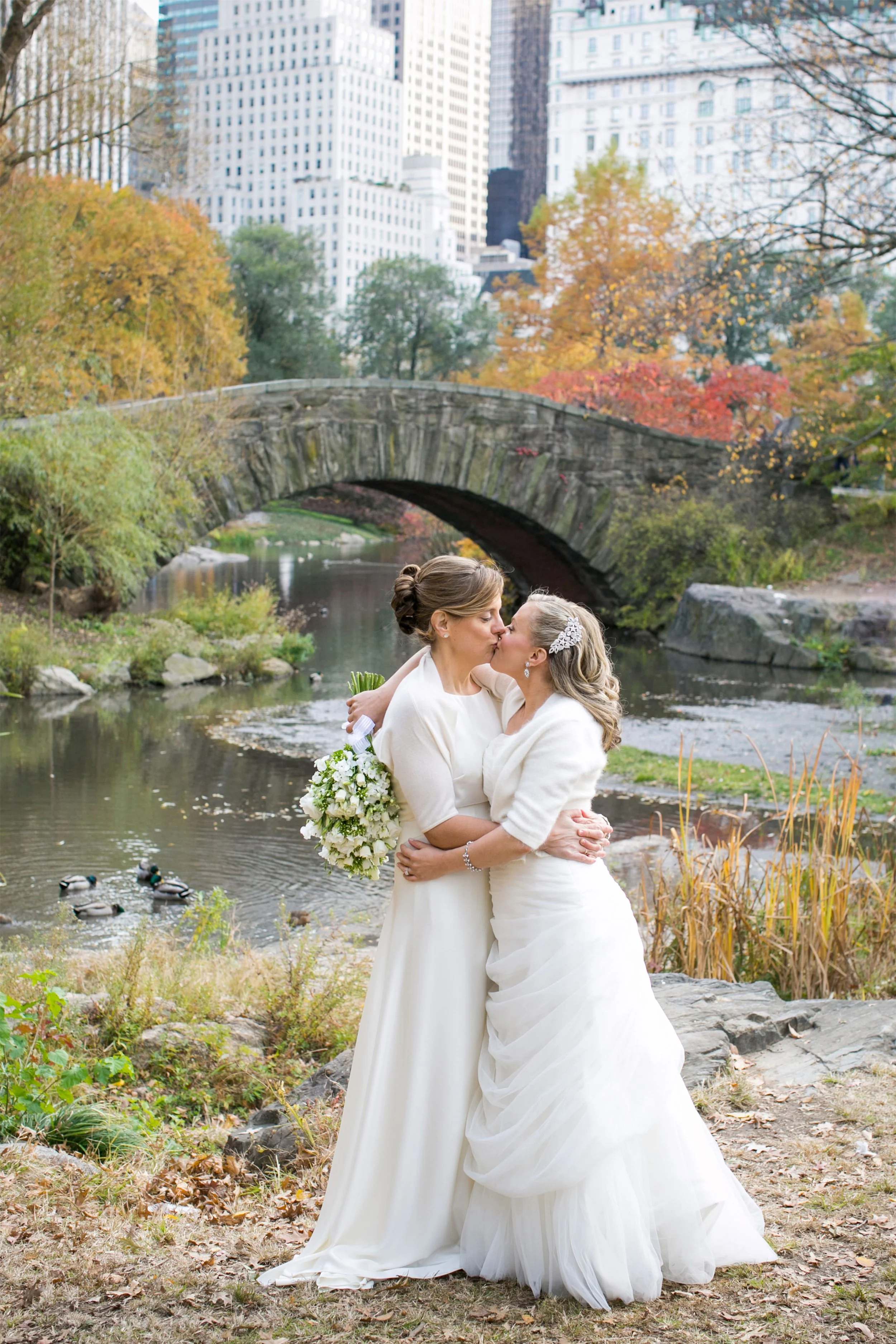 Two women in wedding dresses embracing and kissing in a park with a river, trees with fall foliage, ducks in the water, and city skyscrapers in the background.