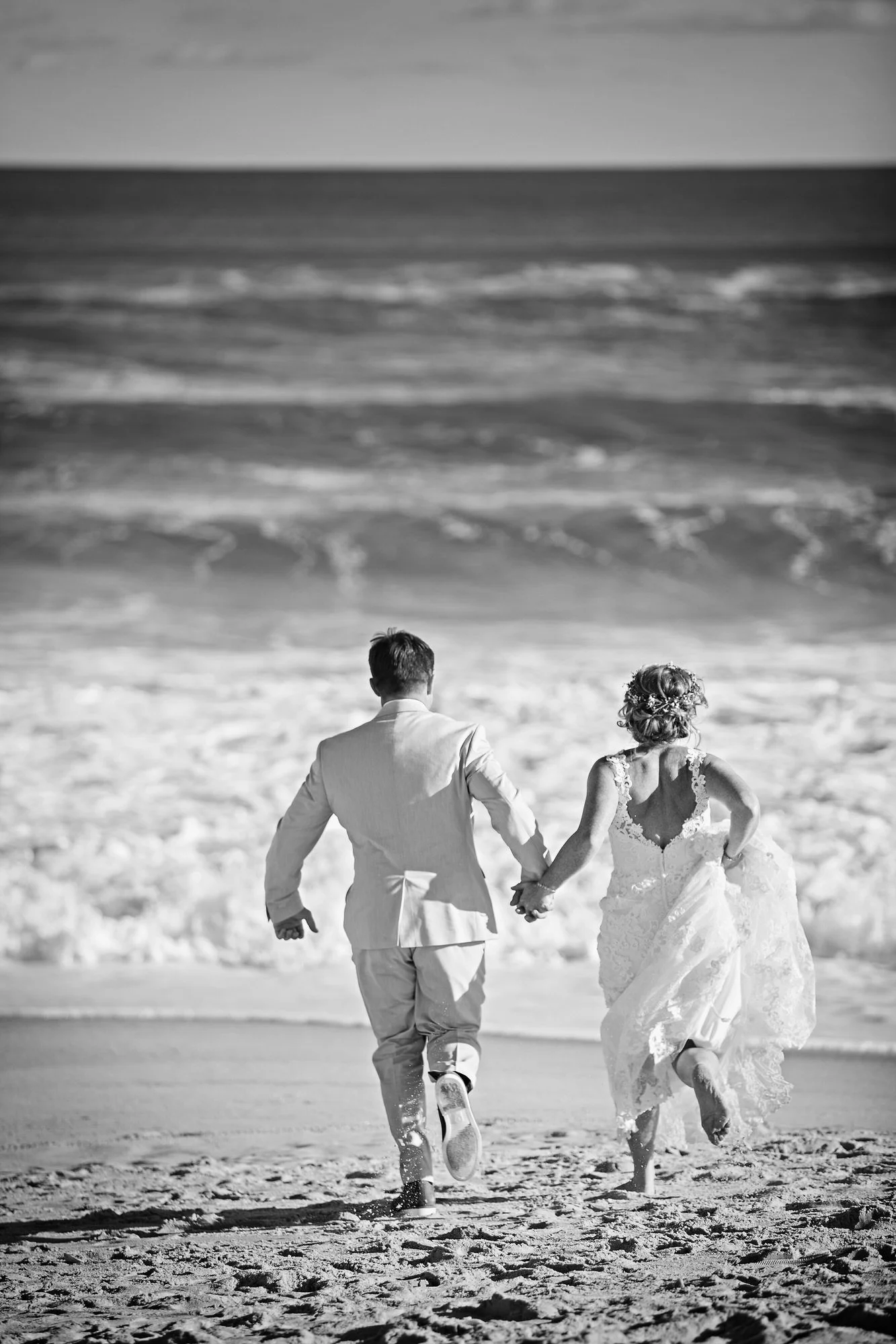 A black and white photograph of a bride and groom running hand in hand on the beach, with waves in the background.