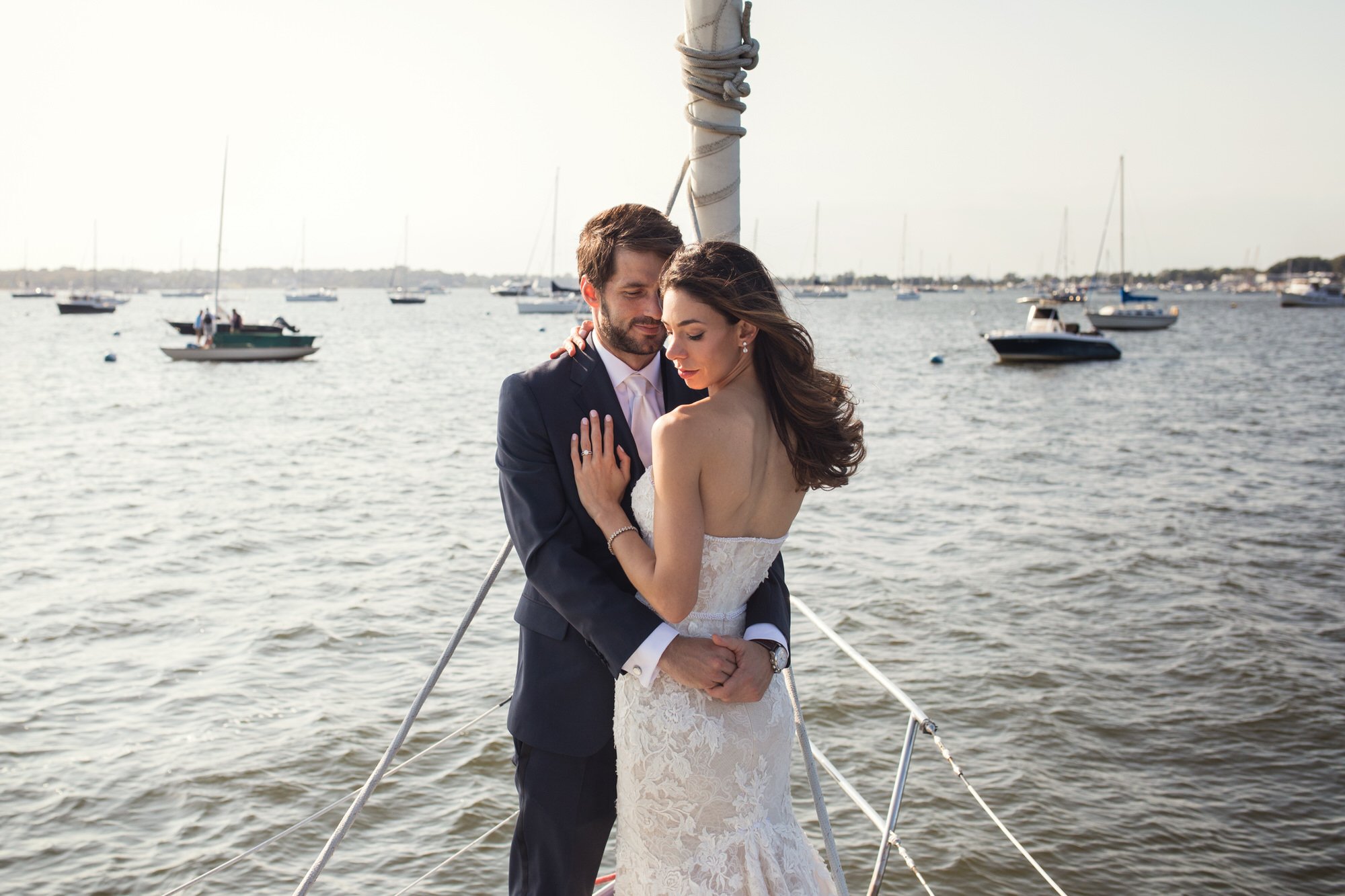 A newlywed couple embracing on a sailboat with a marina and boats in the background.