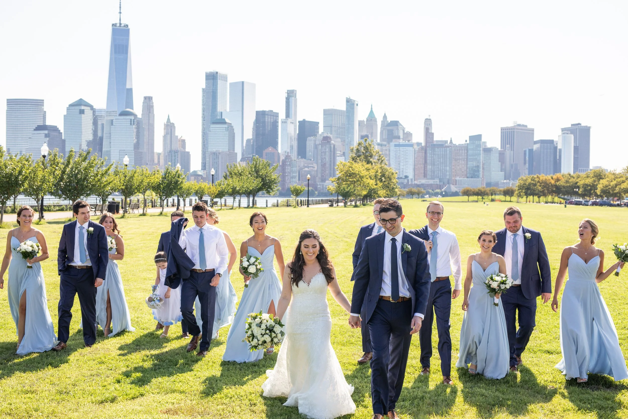 A wedding party walking on a grassy area with New York City skyline in the background, including One World Trade Center, under a sunny sky.