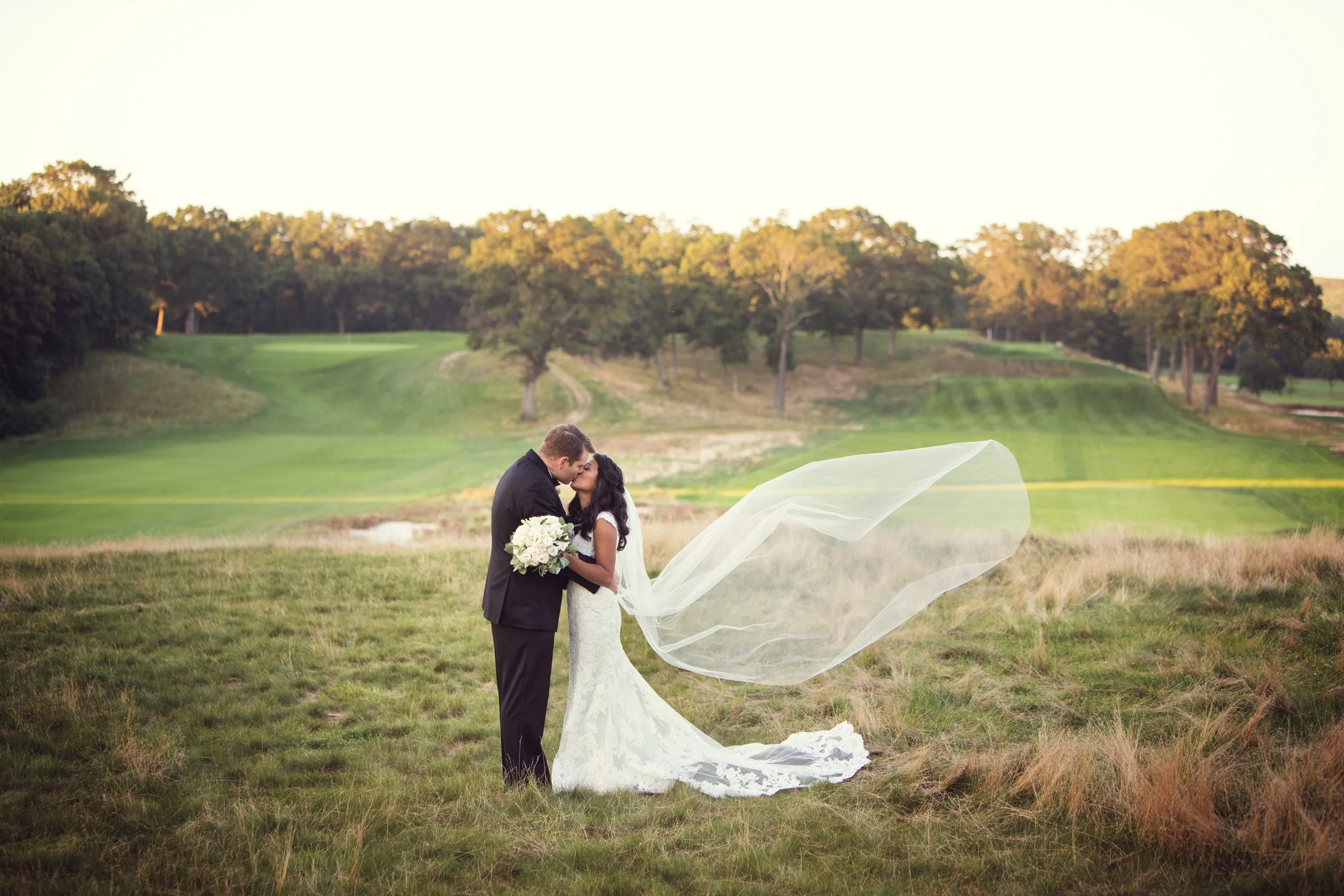 A newlywed couple kissing on a grassy field with a golf course and trees in the background, the bride holding a bouquet of white flowers and her veil flowing in the wind.