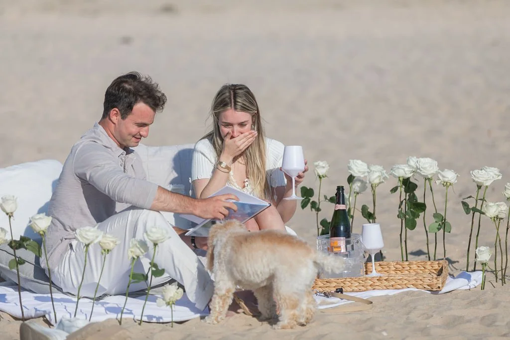 A couple having a picnic on the beach with white roses, a bottle of wine, and a dog.