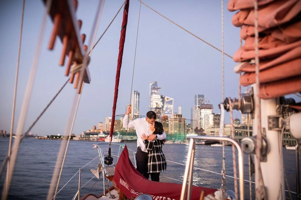 A couple is dancing on the deck of a sailboat with a city skyline in the background at sunset.