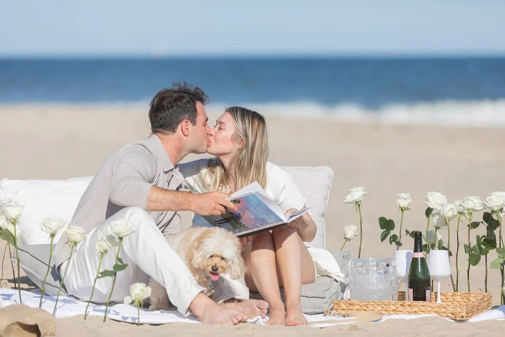 A couple sharing a kiss on a beach blanket, with a small dog, white roses, a bottle of champagne, and a picnic basket nearby.