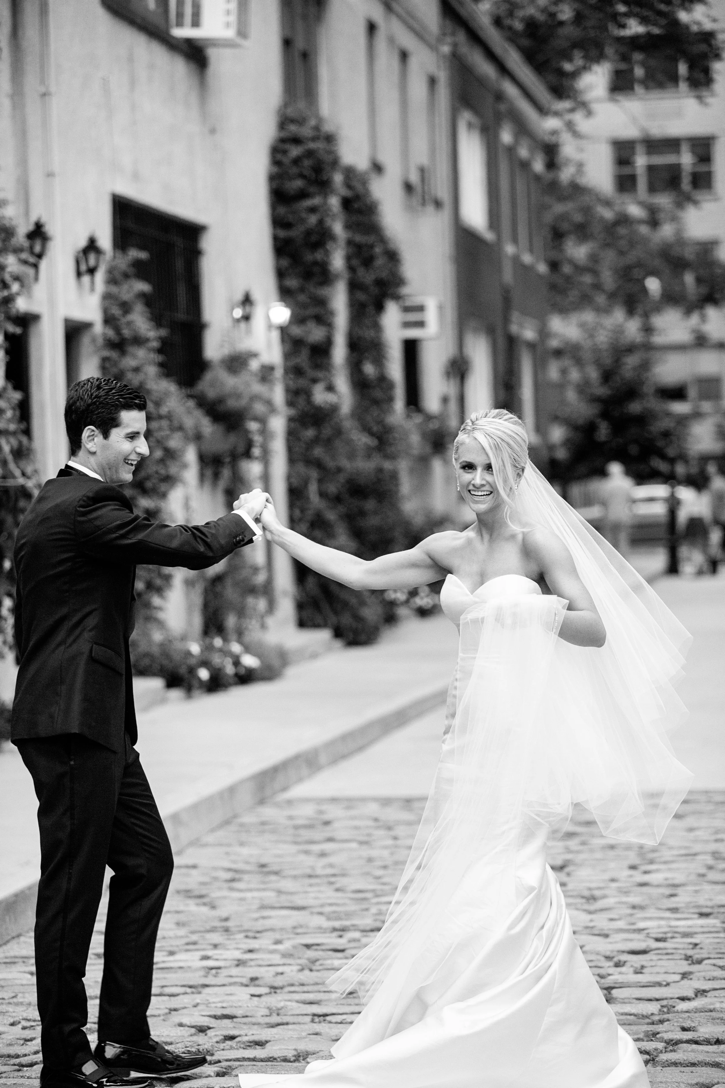 A bride and groom are dancing on a cobblestone street in an urban area, smiling and holding hands, dressed in wedding attire with buildings and trees in the background.