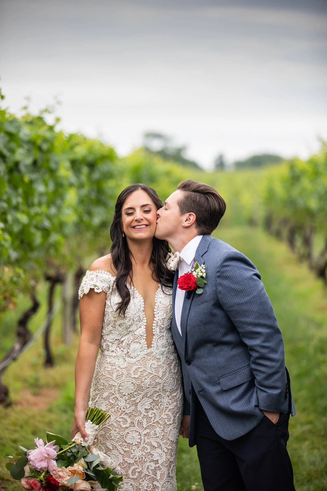 A bride and groom in a vineyard, with the groom kissing the bride on the cheek. The bride is smiling and holding a bouquet, while the groom is dressed in a blue suit with a red flower boutonniere.