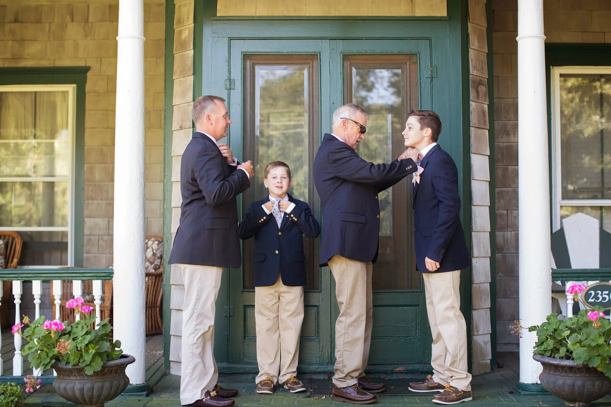 Four males, including a young man, two boys, and an older man, dressed in matching navy blazers and khaki pants, standing on the porch of a house with green door, preparing for a formal event.