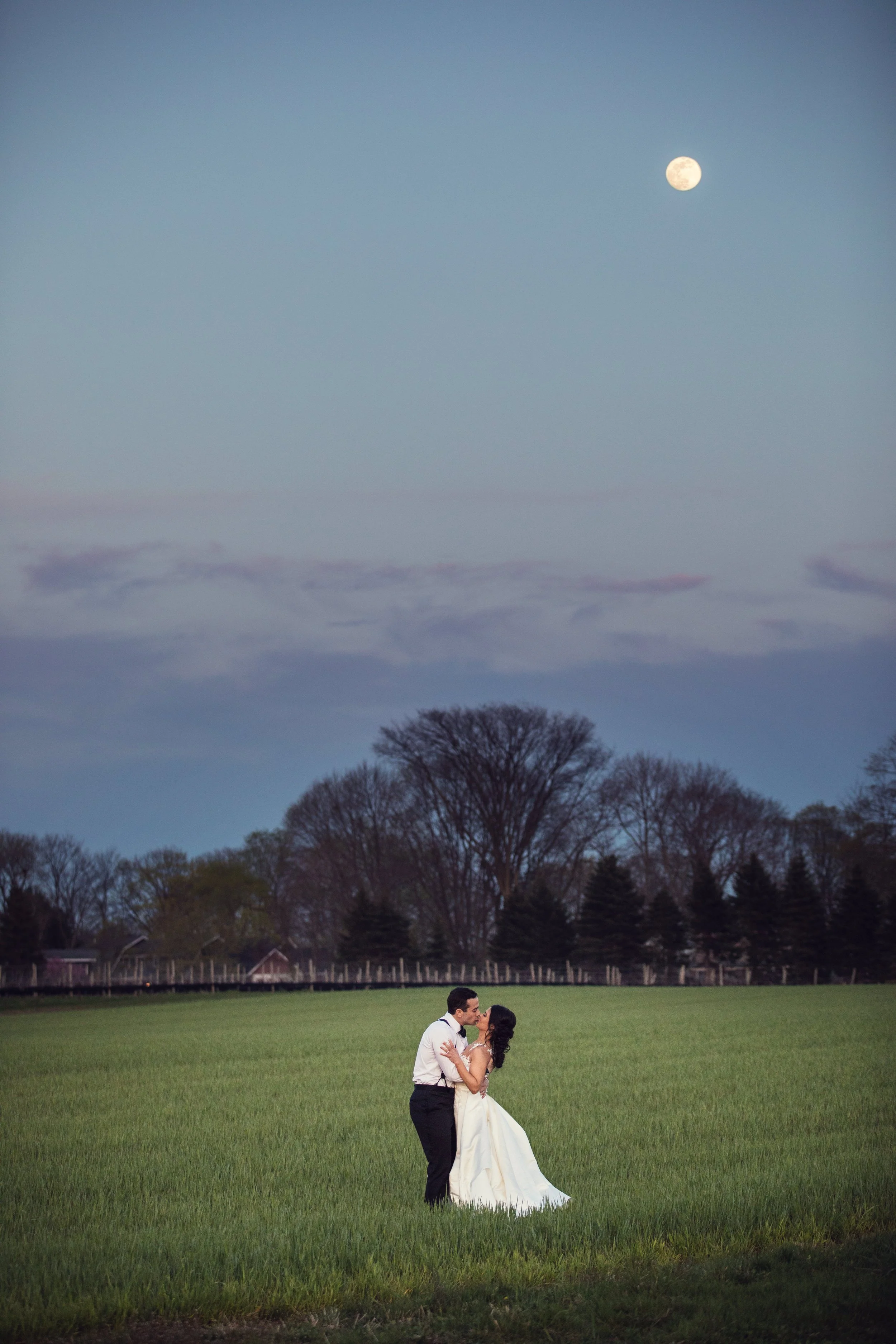 A couple dressed in wedding attire dancing in a green field at dusk with the moon visible in the sky and trees in the background.