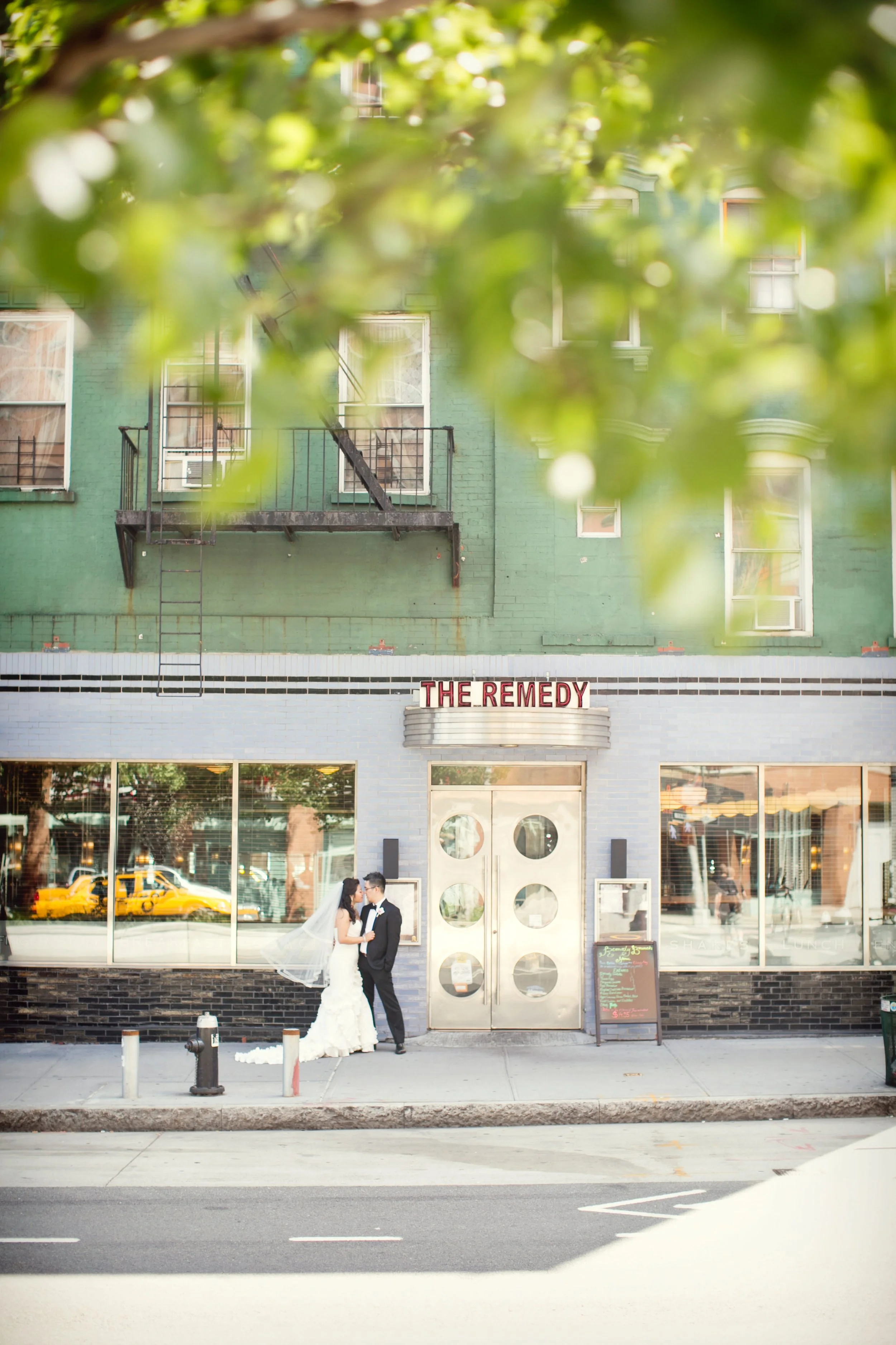 A bride and groom in wedding attire standing outside a building called 'The Remedy', with a couple of large glass windows reflecting a yellow taxi, and a blackboard menu on the sidewalk.