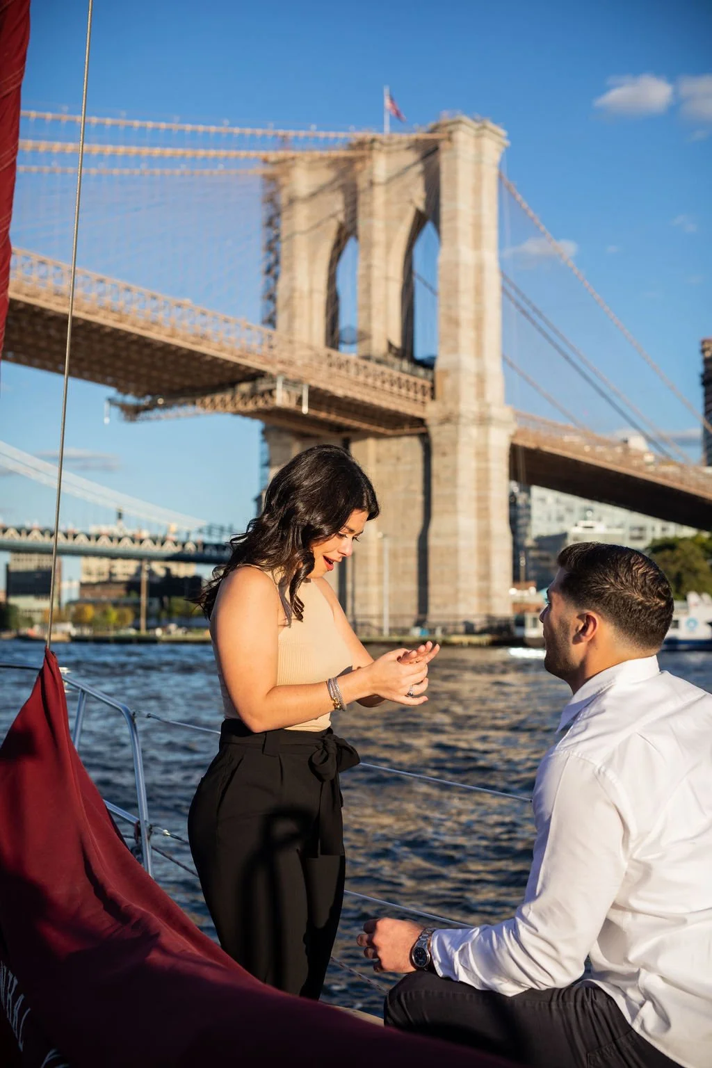 A couple on a boat near the Brooklyn Bridge in New York City, with the woman proposing to the man during sunset.