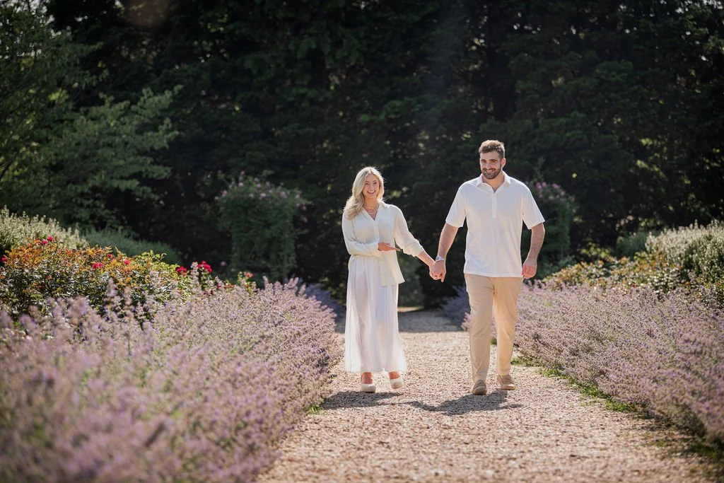 A couple holding hands and walking on a gravel path through a garden with blooming flowers, surrounded by greenery and trees.