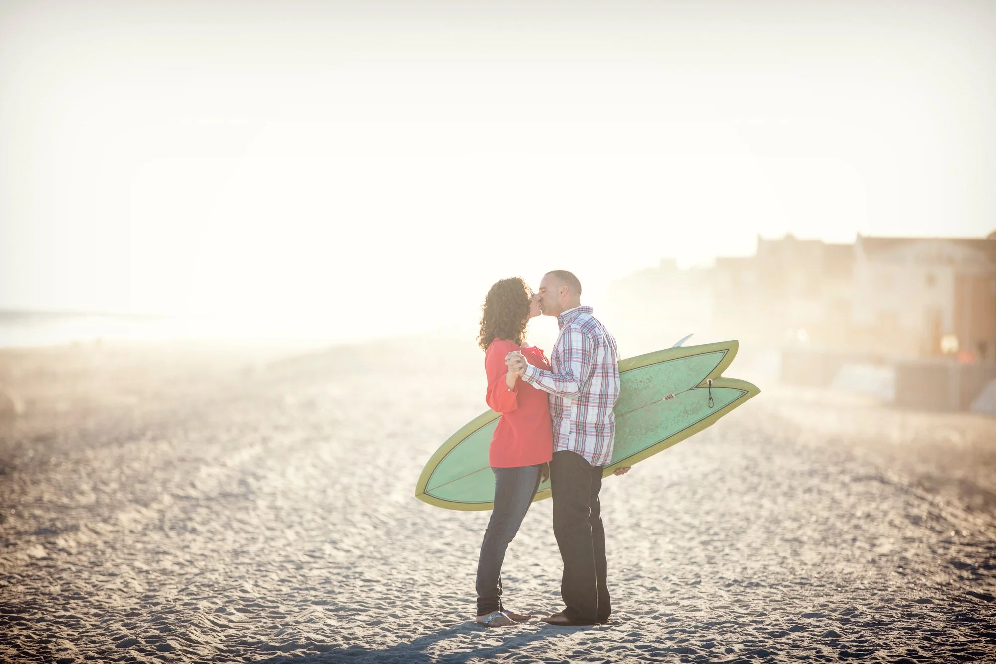 A couple kissing on the beach, holding hands with a surfboard.