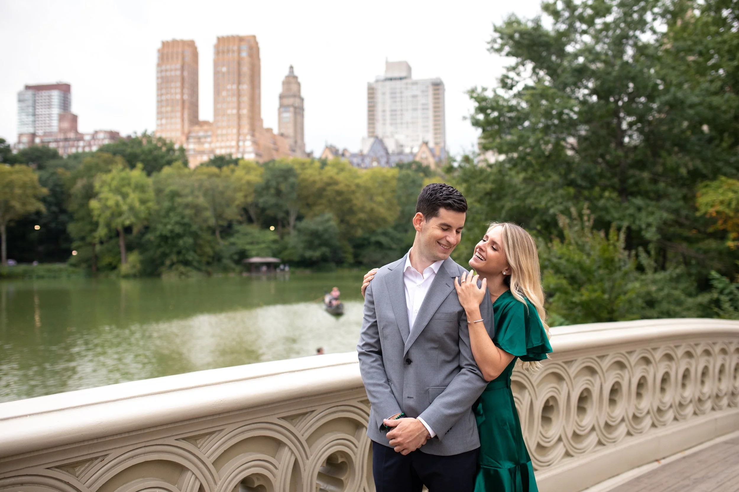 A happy couple sharing a joyful moment by a river in a park, with city buildings and greenery in the background.