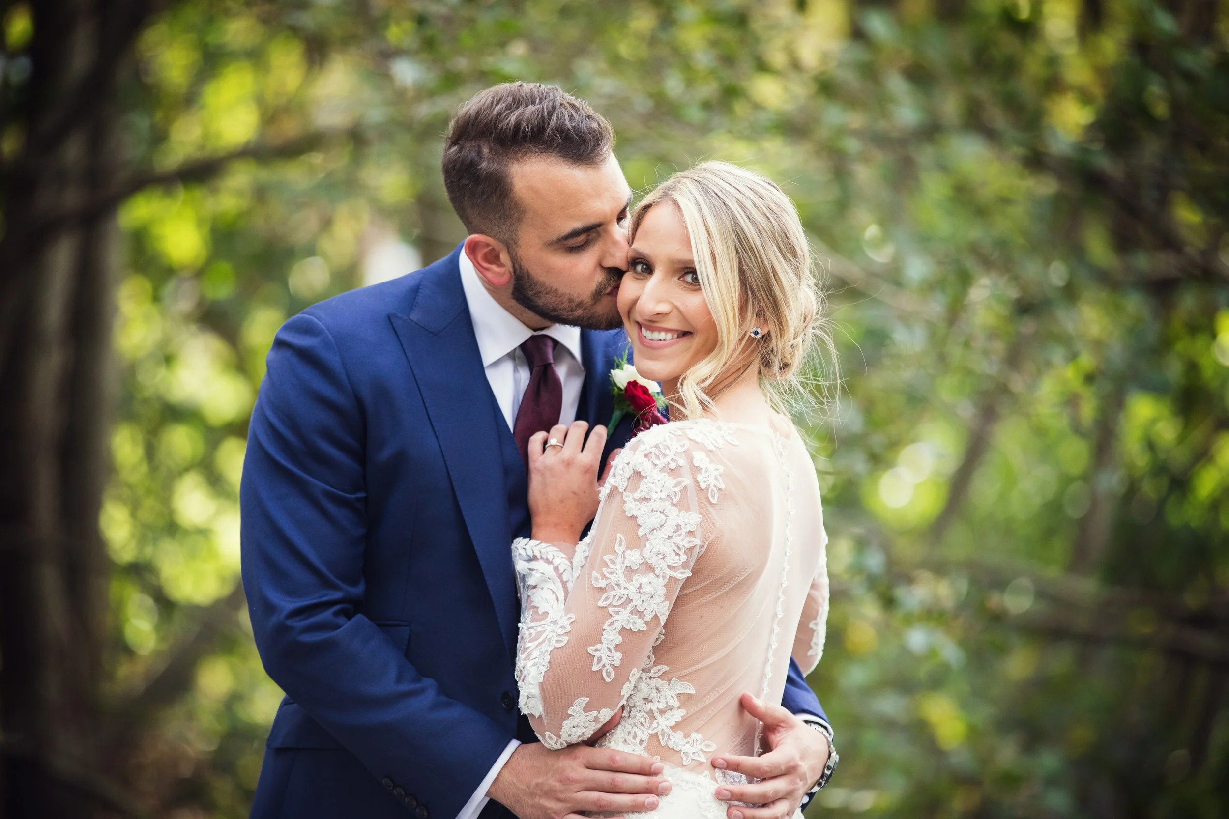 A newlywed couple in wedding attire embraces outdoors with a green, blurred forest background. The groom, dressed in a blue suit, kisses the bride on the cheek. The bride, in a lace wedding dress, smiles at the camera.