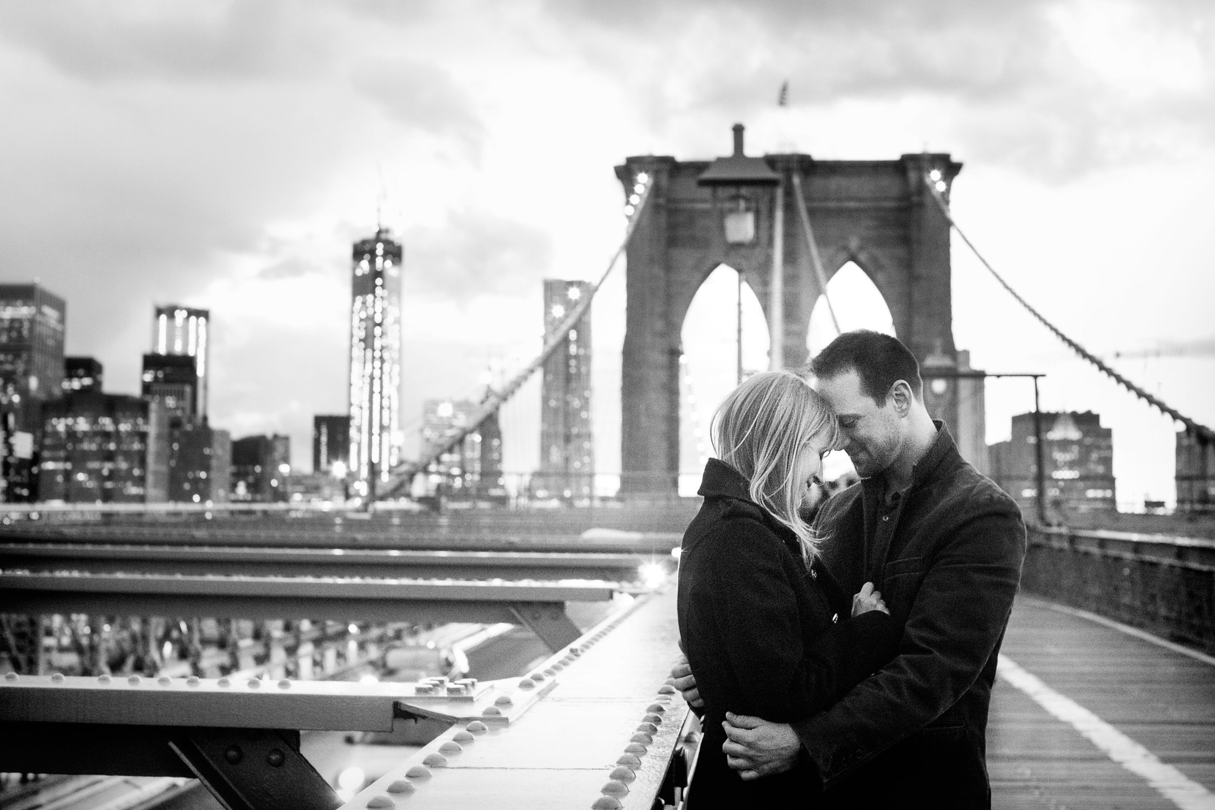 A black and white photo of a couple embracing on the Brooklyn Bridge, with the Manhattan skyline and tall buildings in the background.
