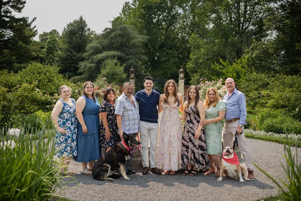 A group of nine people standing on a gravel path in a lush garden, with two dogs, one sitting and one standing, surrounded by greenery and flowering bushes.