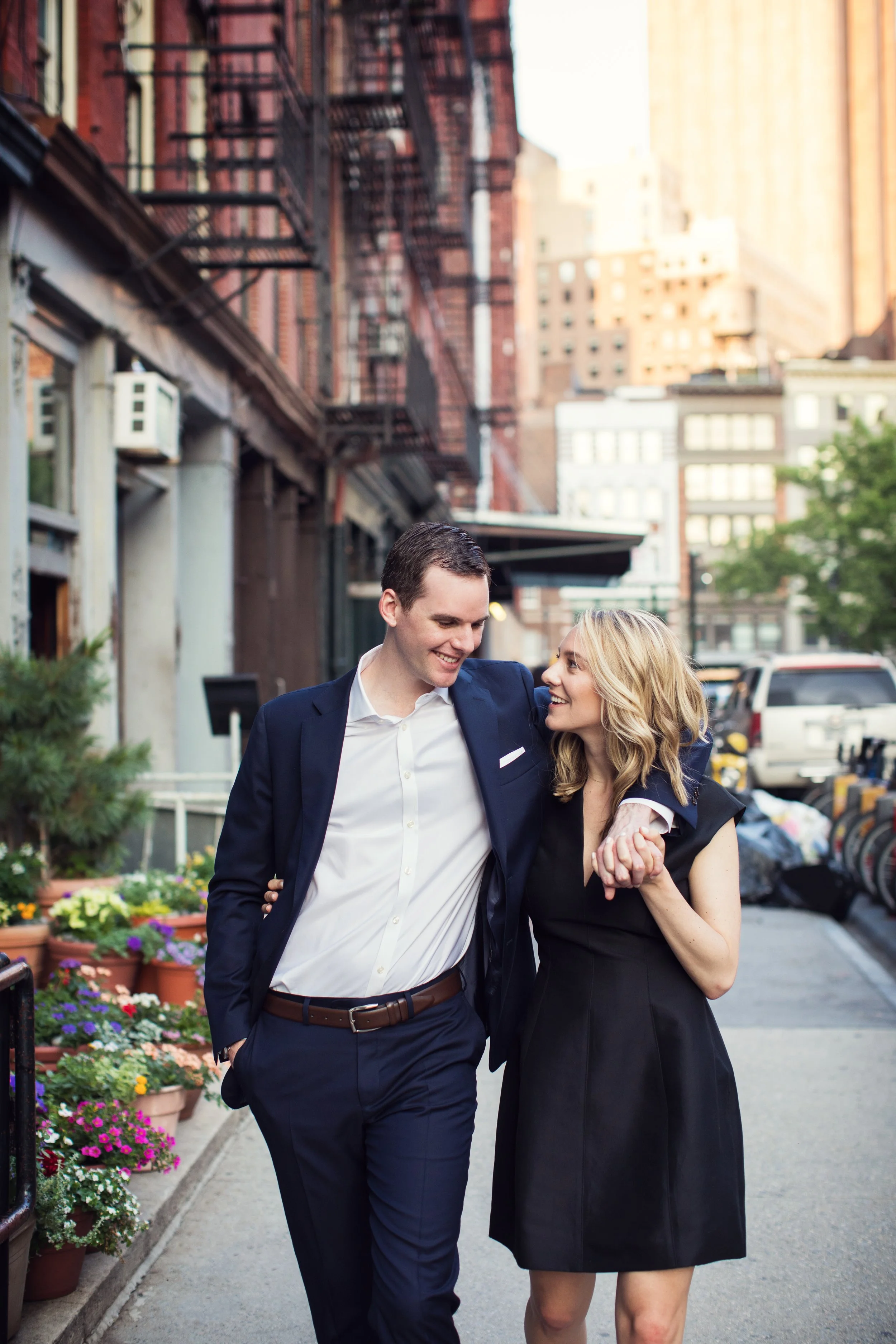 A happy man and woman walking together on a city sidewalk, smiling at each other, with buildings and parked cars in the background, and potted flowers lining the sidewalk.