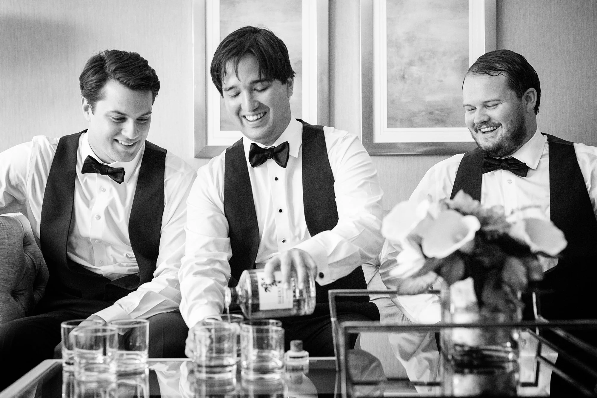Three men in tuxedos pouring drinks and smiling at a table with glasses and a floral arrangement.
