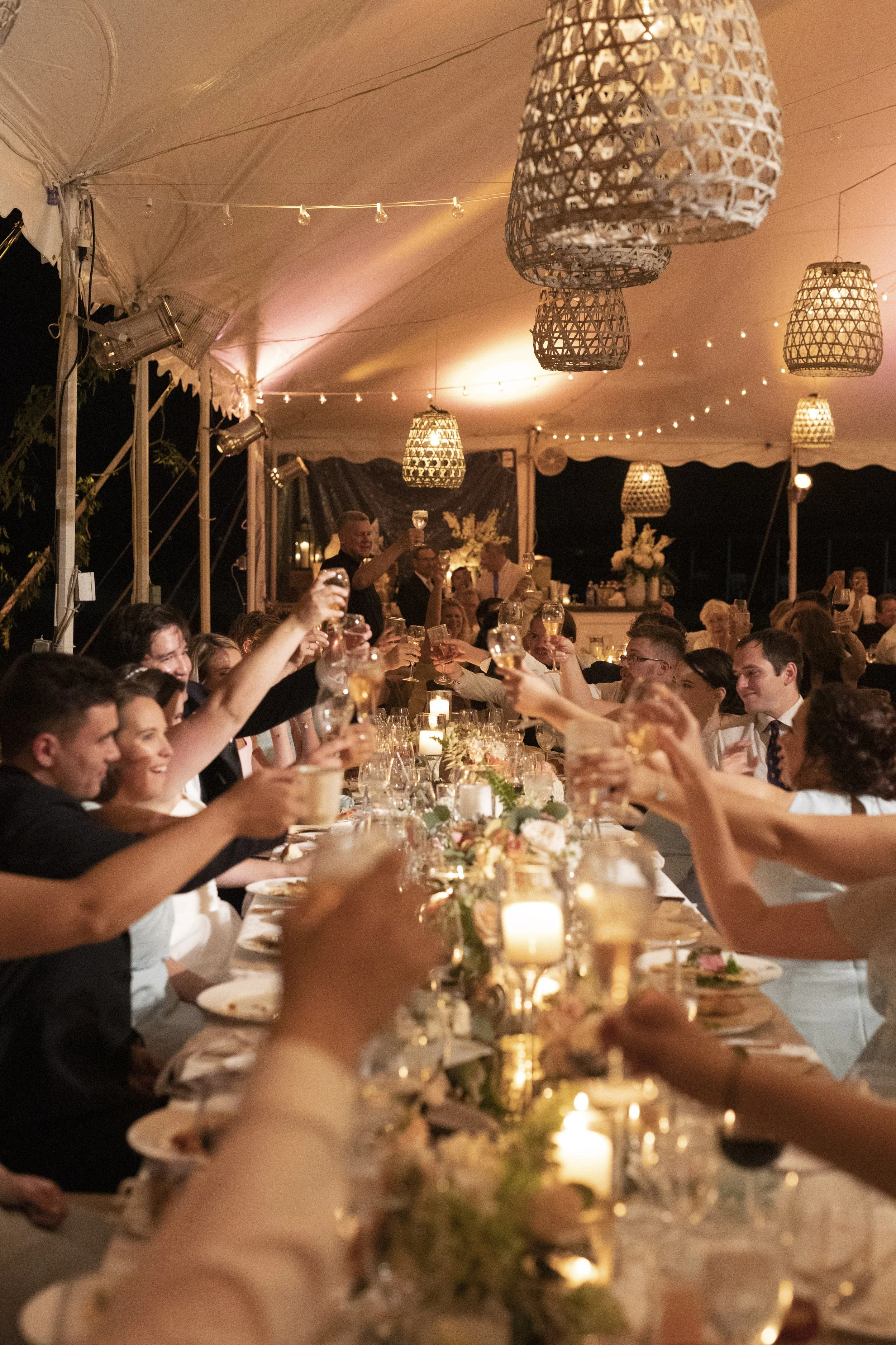 People raising glasses in toast at a long dinner table under string lights and woven pendant lamps during an evening celebration.