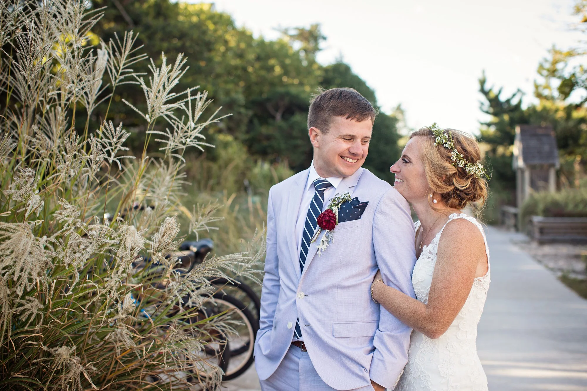 A bride and groom smiling at each other outdoors during their wedding, with plants and a pathway in the background.