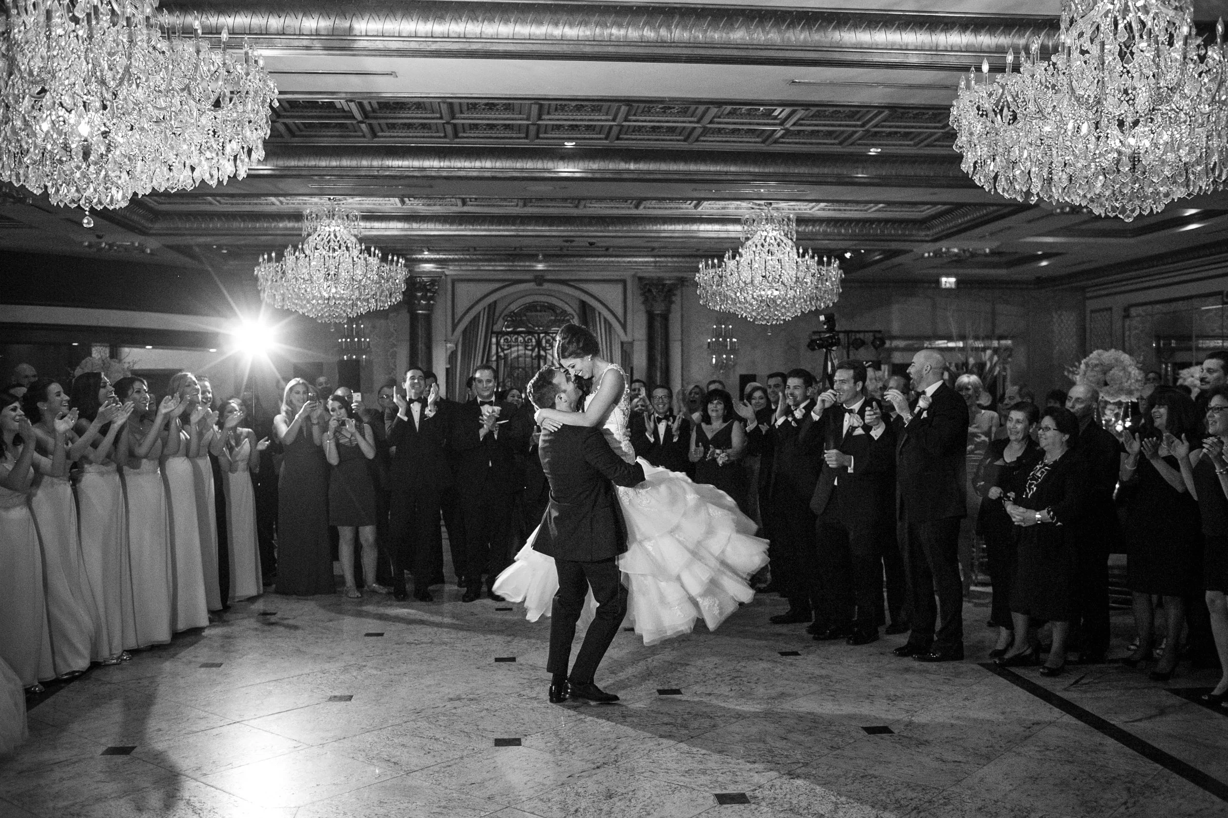 A wedding reception with a bride and groom dancing in the center, surrounded by guests clapping and cheering in an elegant ballroom with chandeliers.