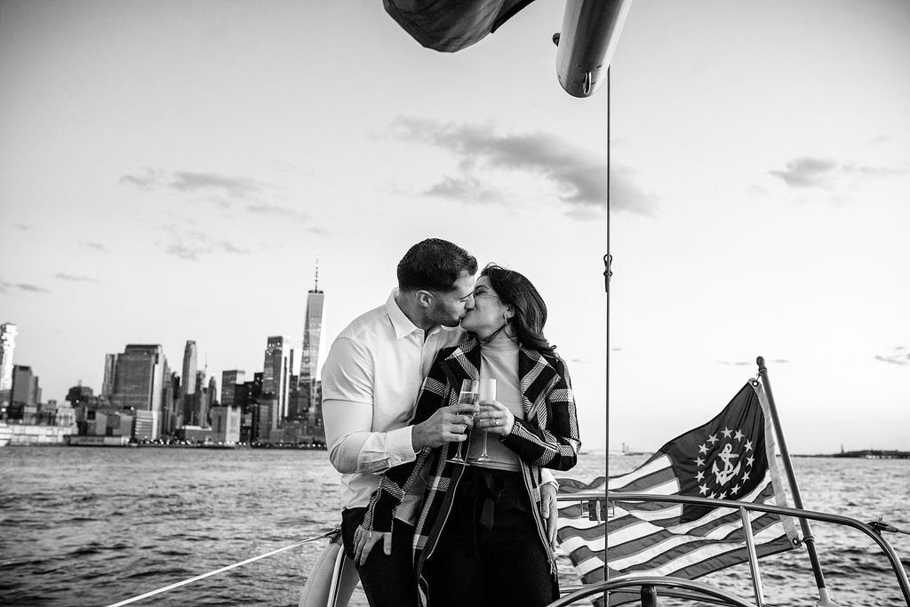 A couple kissing on a boat with the New York City skyline in the background, holding champagne glasses. The boat has an American flag with anchor design.