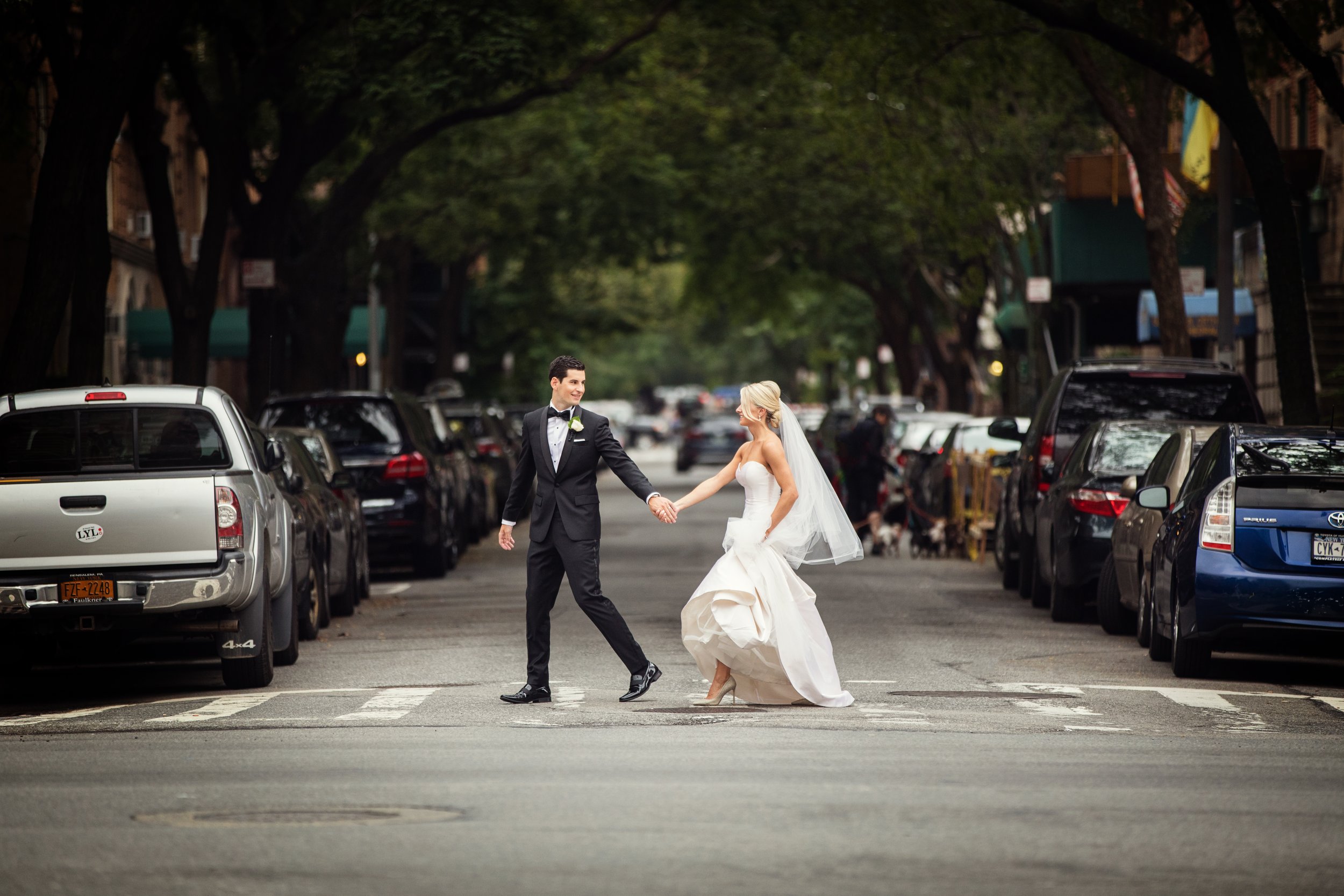 A newlywed couple walking hand in hand across a city street with parked cars on both sides, green trees overhead, and a blurred background of more cars and pedestrians.