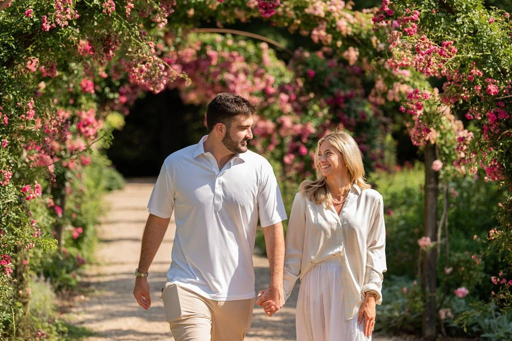 A happy couple walking hand in hand through a garden archway covered with pink and white flowers.