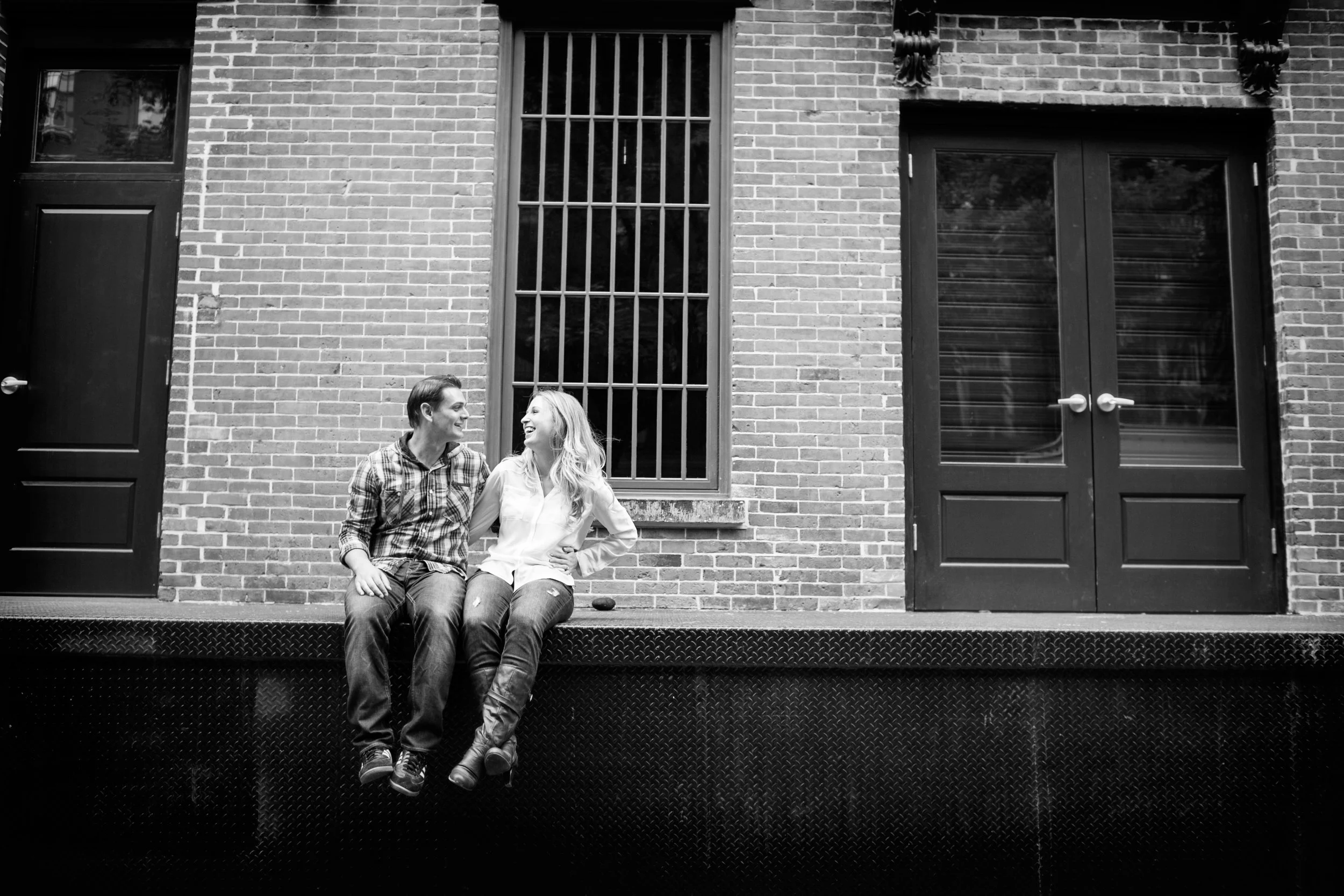 A black-and-white photo of a young man and woman sitting on a platform outside a brick building, smiling and looking at each other.