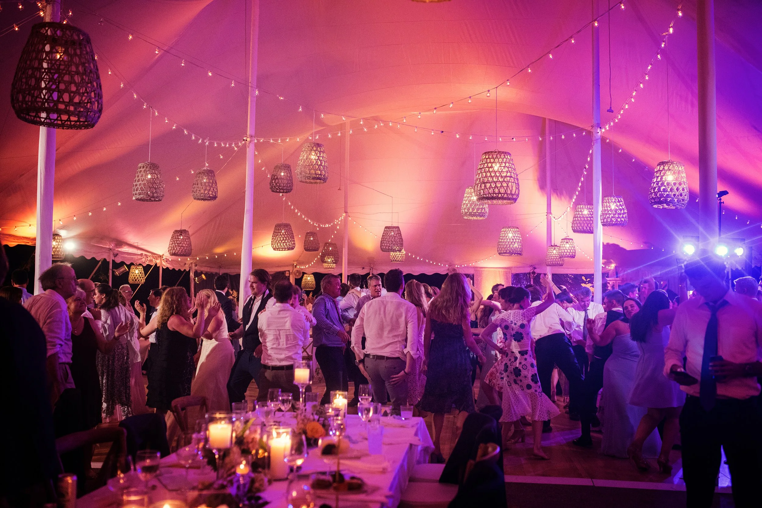 People dancing and socializing under hanging lanterns and string lights at a lively party or celebration in a large tent with purple and pink lighting.