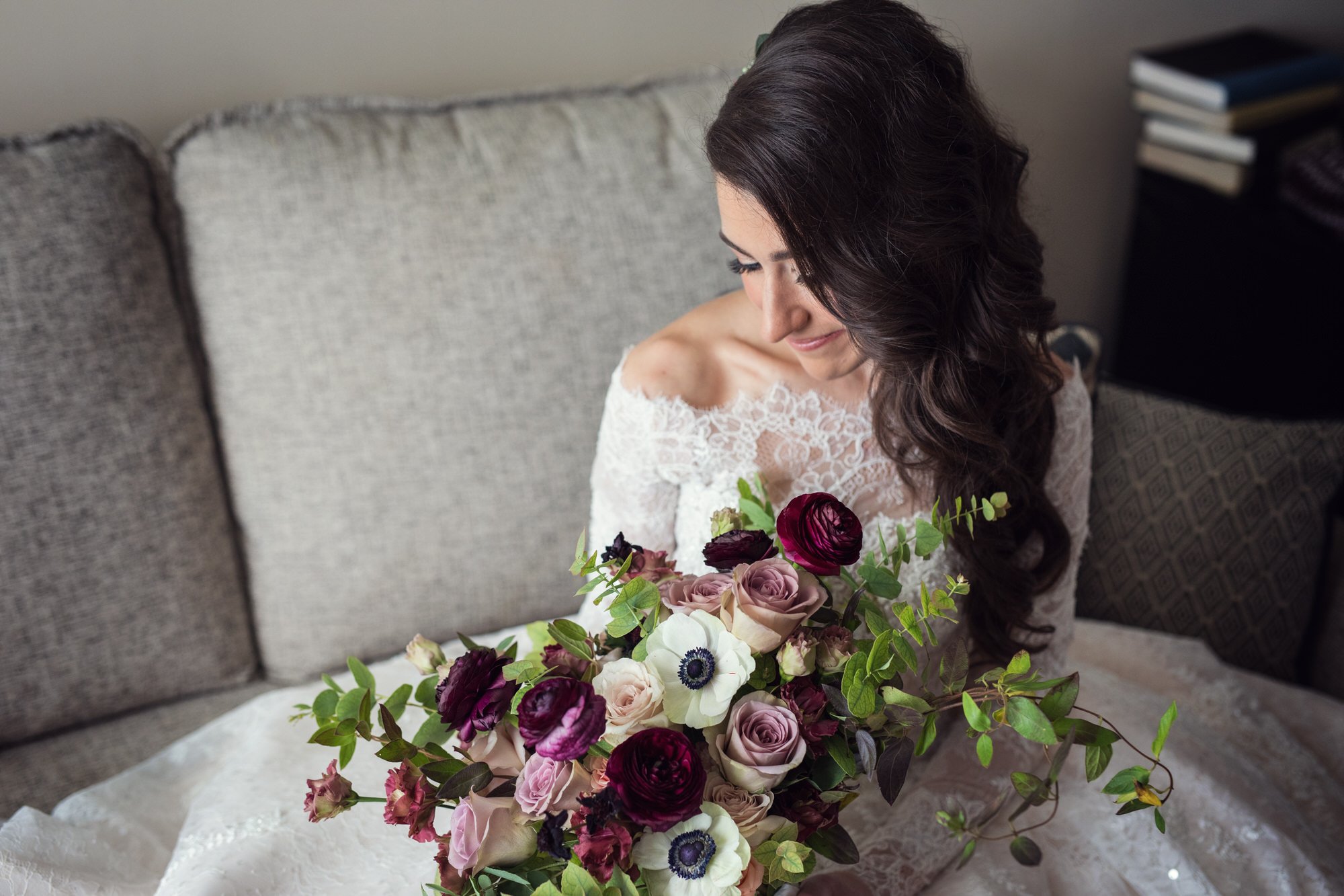 A woman with long, dark, wavy hair in a white lace dress looks down at a large bouquet of pink, purple, and white flowers with green leaves, sitting on her lap in a cozy room.