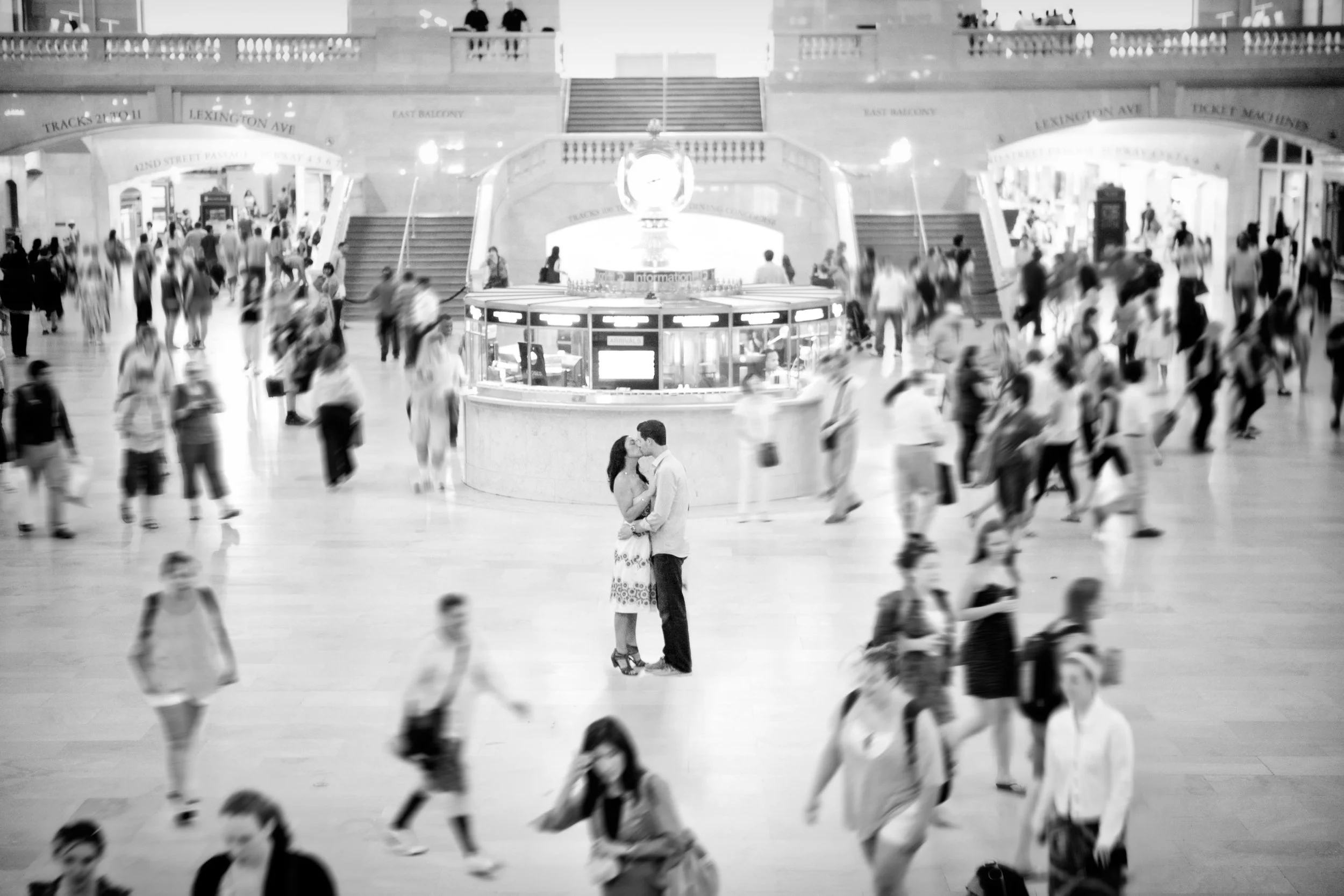 Black and white image of a busy train station with many people walking around. In the center, a couple is kissing, surrounded by a crowd of commuters.