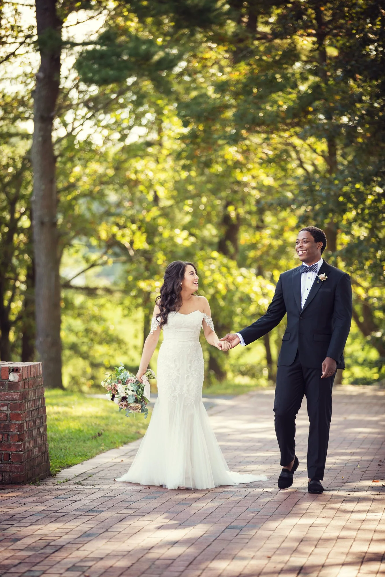 A bride and groom holding hands and smiling while walking outdoors in a park-like setting with trees and greenery in the background. The bride is wearing a white wedding dress and holding a bouquet of flowers, and the groom is dressed in a black tuxe