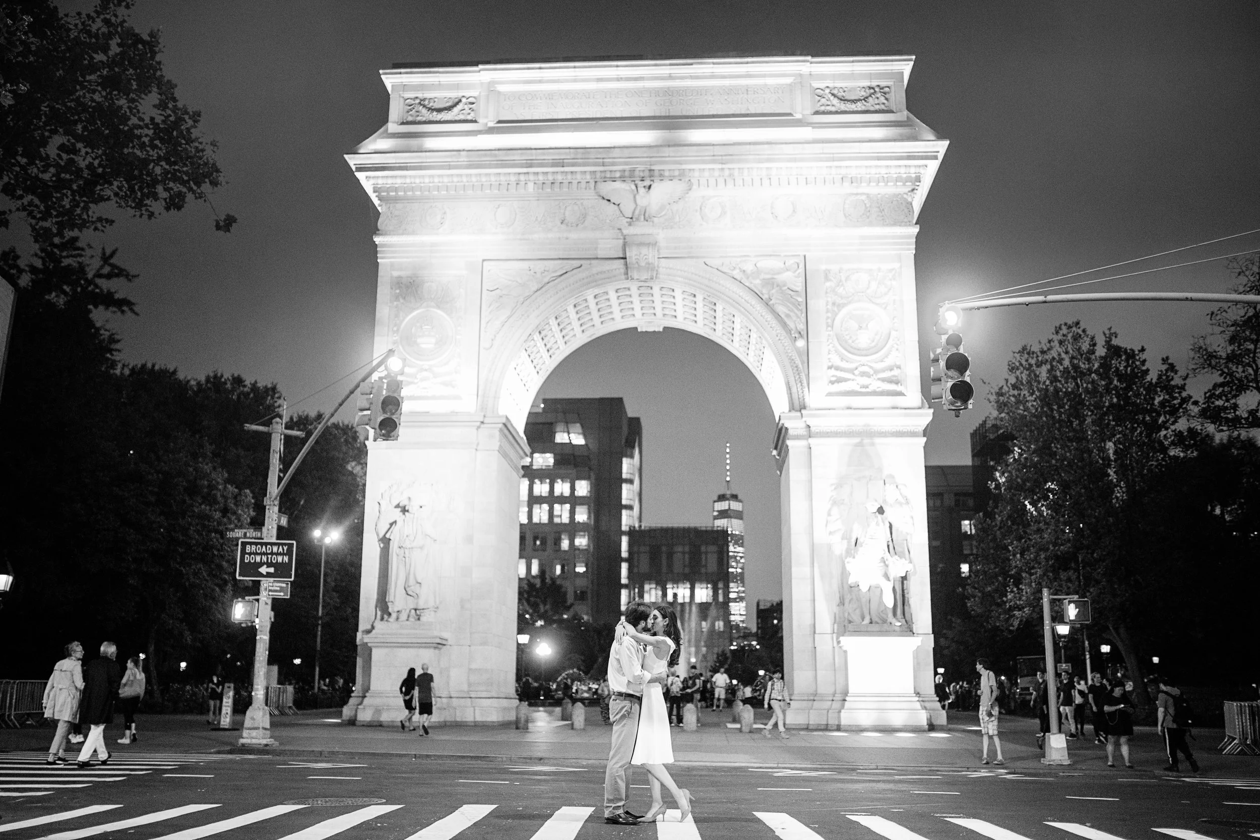 Couple kissing under the Washington Square Arch at night, with people walking in the background