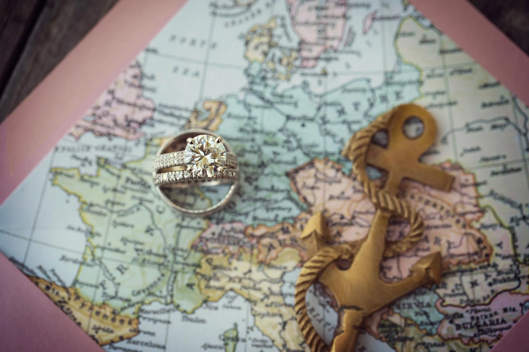 A diamond engagement ring with a large round center stone and a band encrusted with smaller diamonds, placed on a map of Europe. Next to the ring is a gold-colored decorative anchor with a rope design.