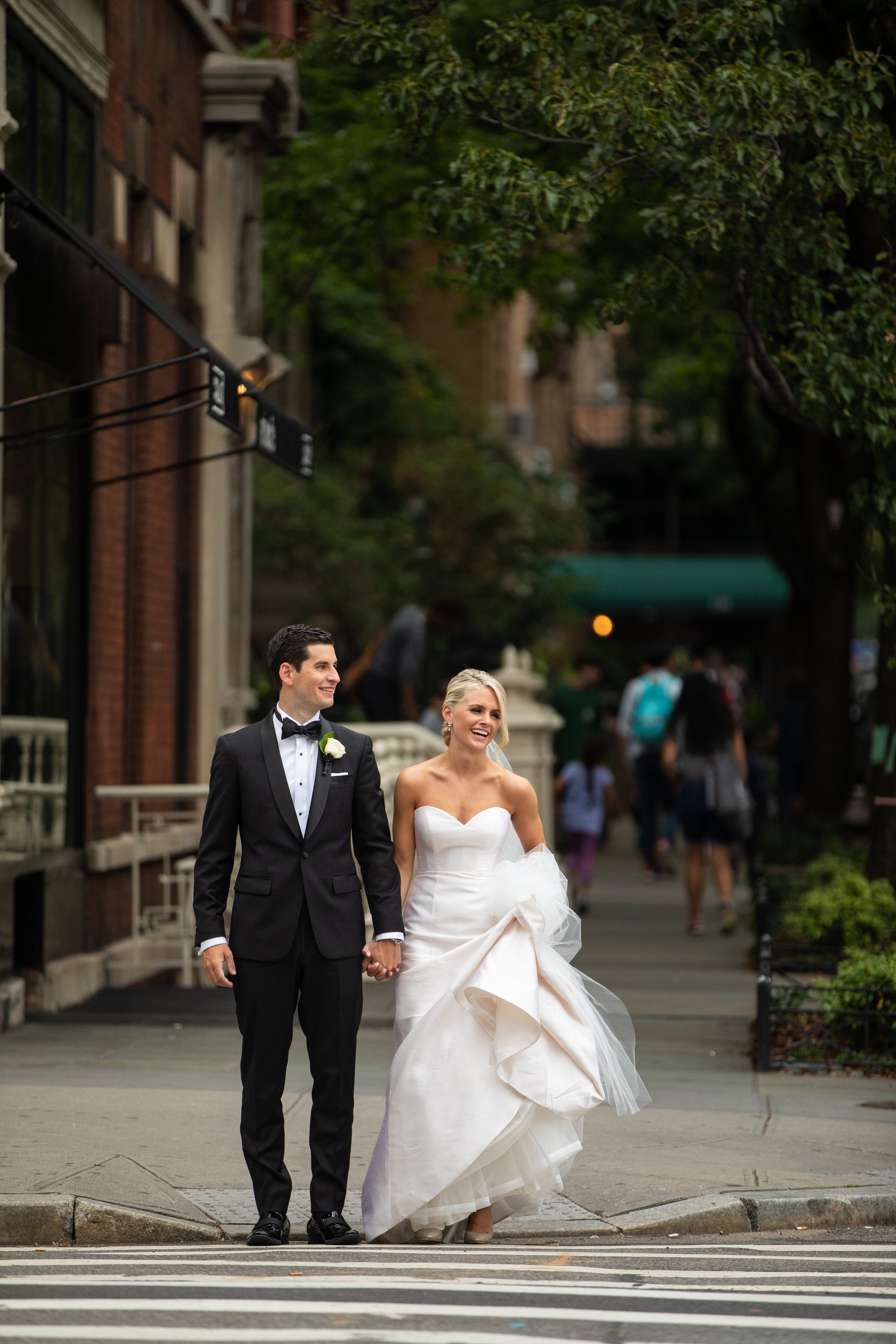 A newlywed couple, dressed in wedding attire, walking hand-in-hand across a city street, smiling and enjoying their moment.
