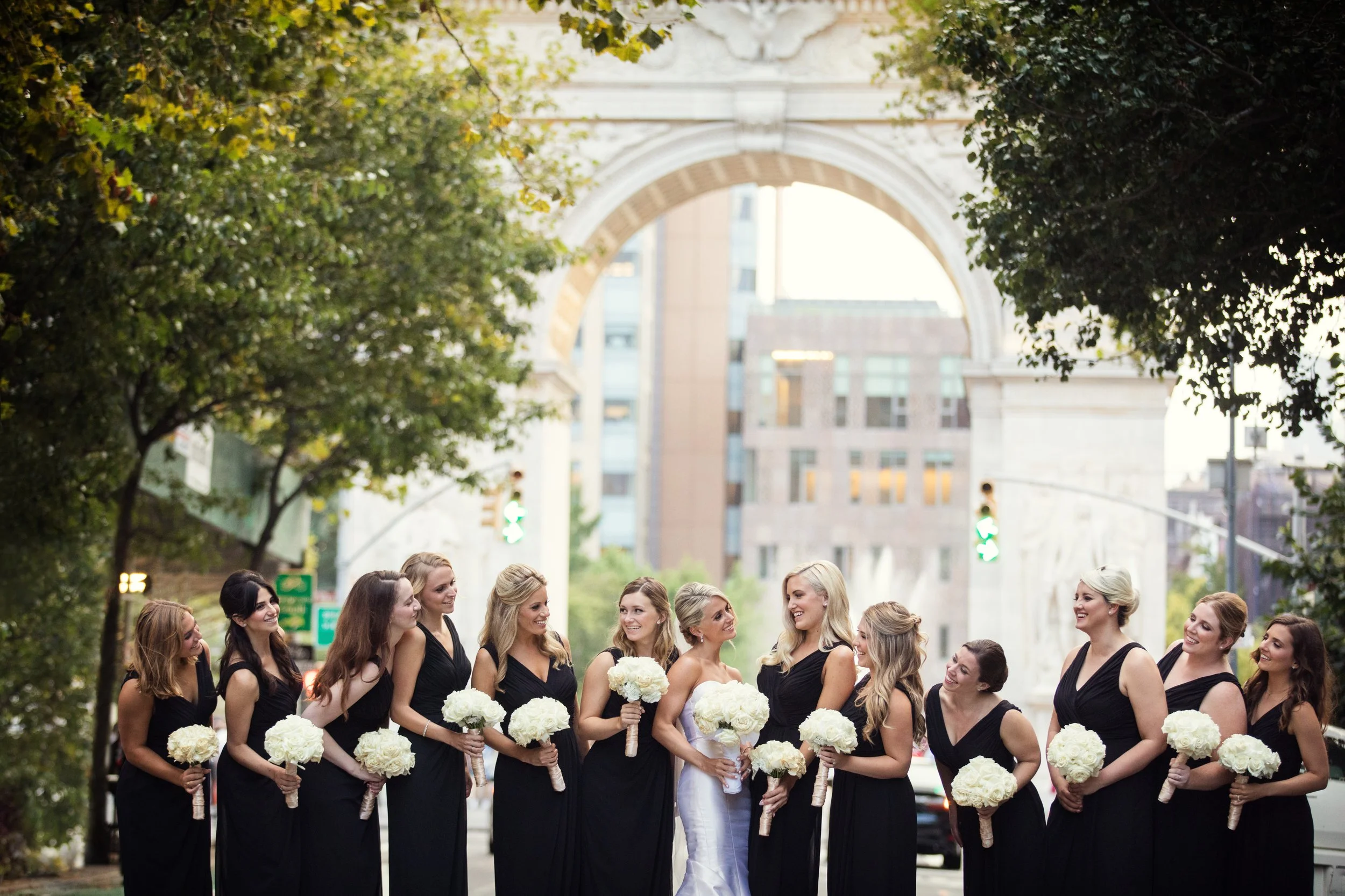 A group of women in black dresses holding white bouquets, standing on a city street with trees and a large arch in the background.
