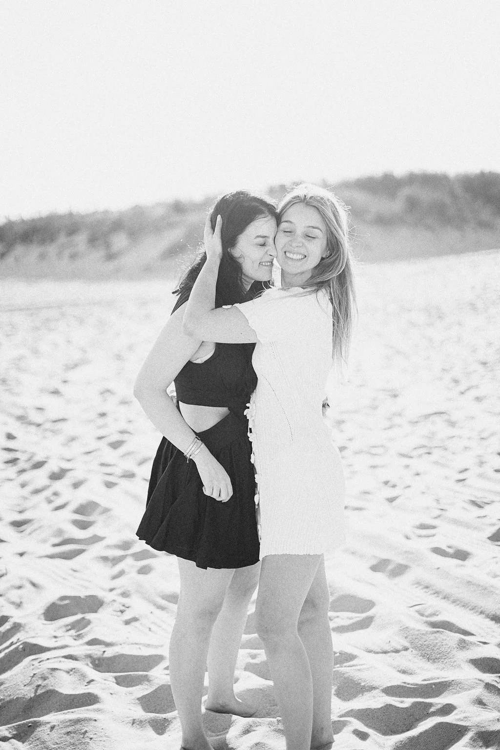Two women are smiling and embracing each other on a sandy beach. One woman has dark hair and is wearing a black dress, while the other woman has blonde hair and is wearing a white dress. The background features the beach and distant dunes.