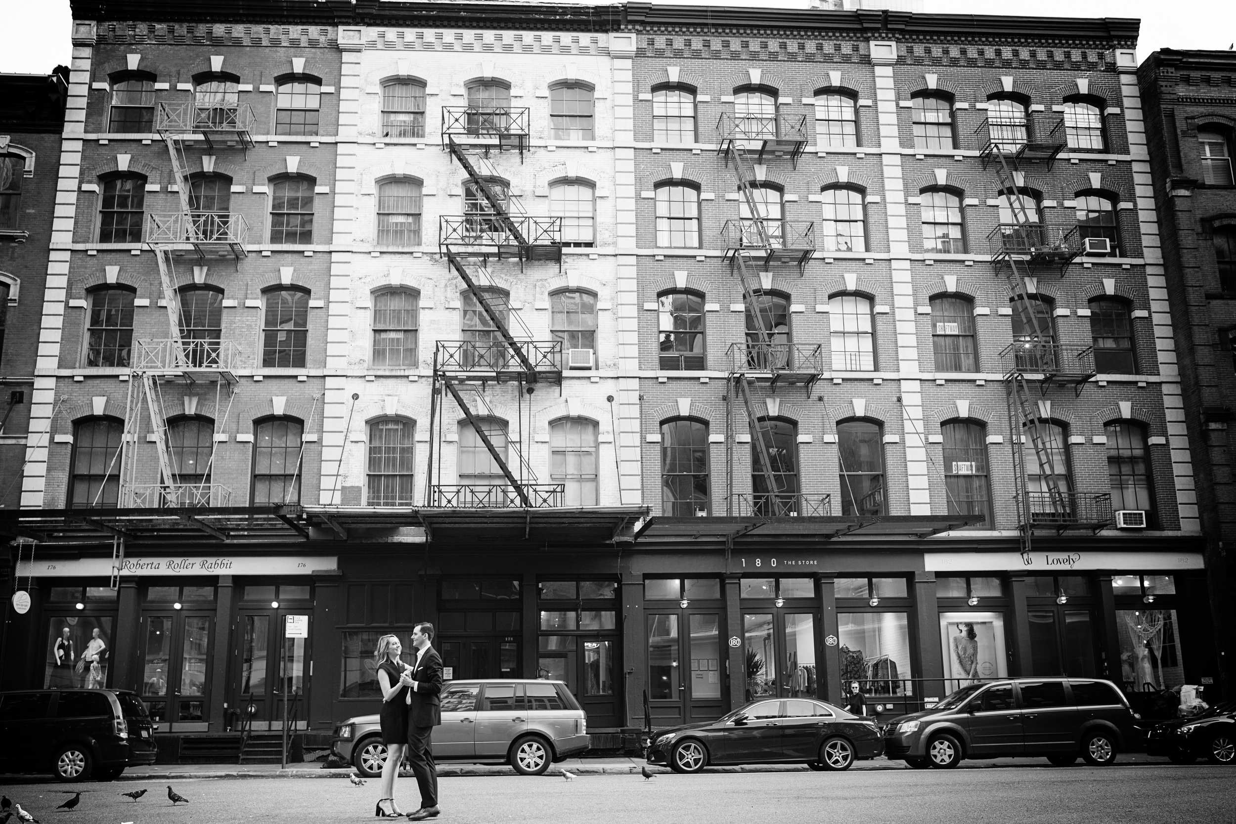 A black and white photo of a city street scene with a building in the background featuring fire escape stairs. In the foreground, a couple dressed formally is dancing or talking romantically near parked cars, with pigeons on the street and a person s