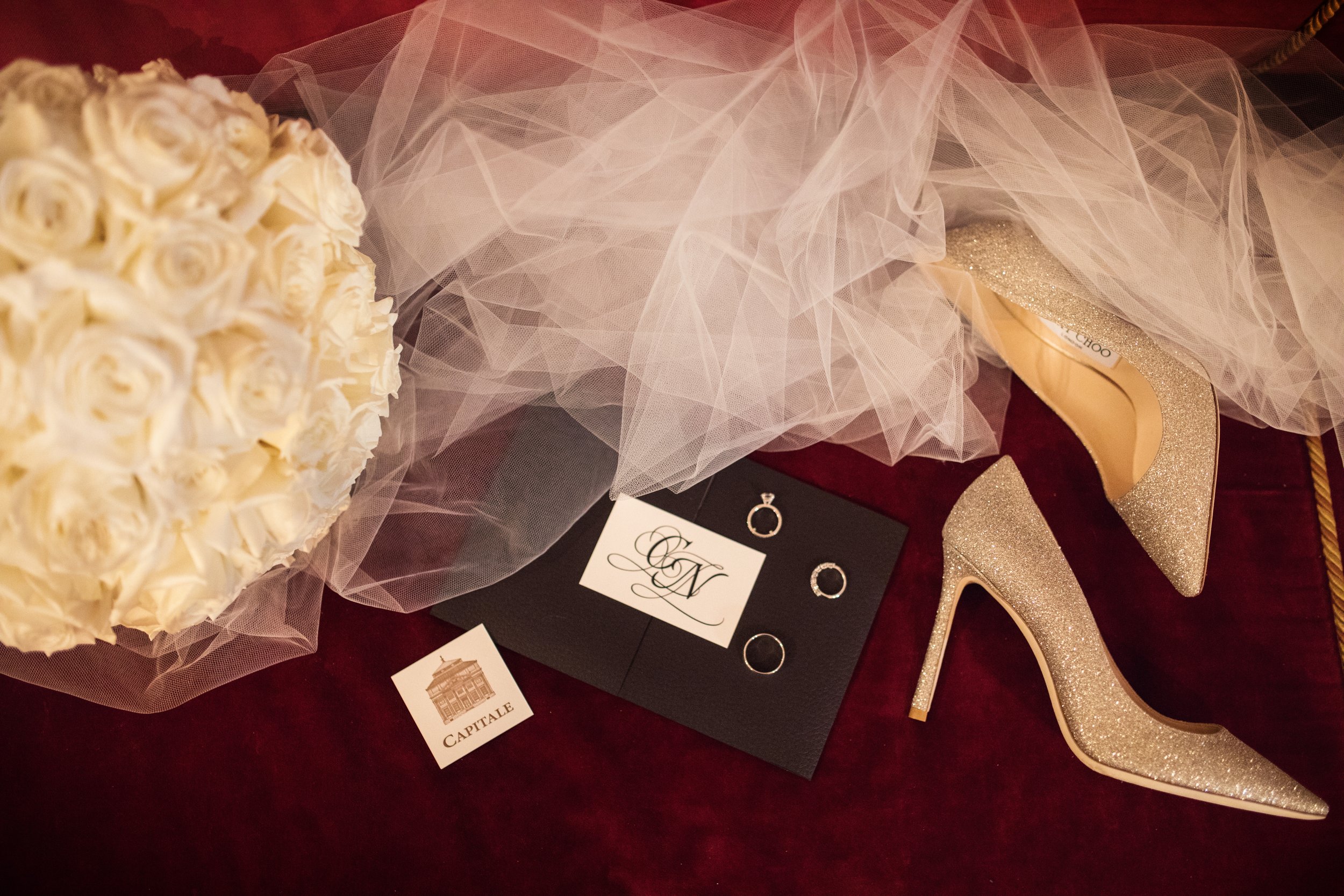 A bouquet of white roses, a pair of gold glitter high heels, a black card with jewelry including rings and a ring box, and a strand of white tulle fabric are arranged on a red surface, likely for a wedding.