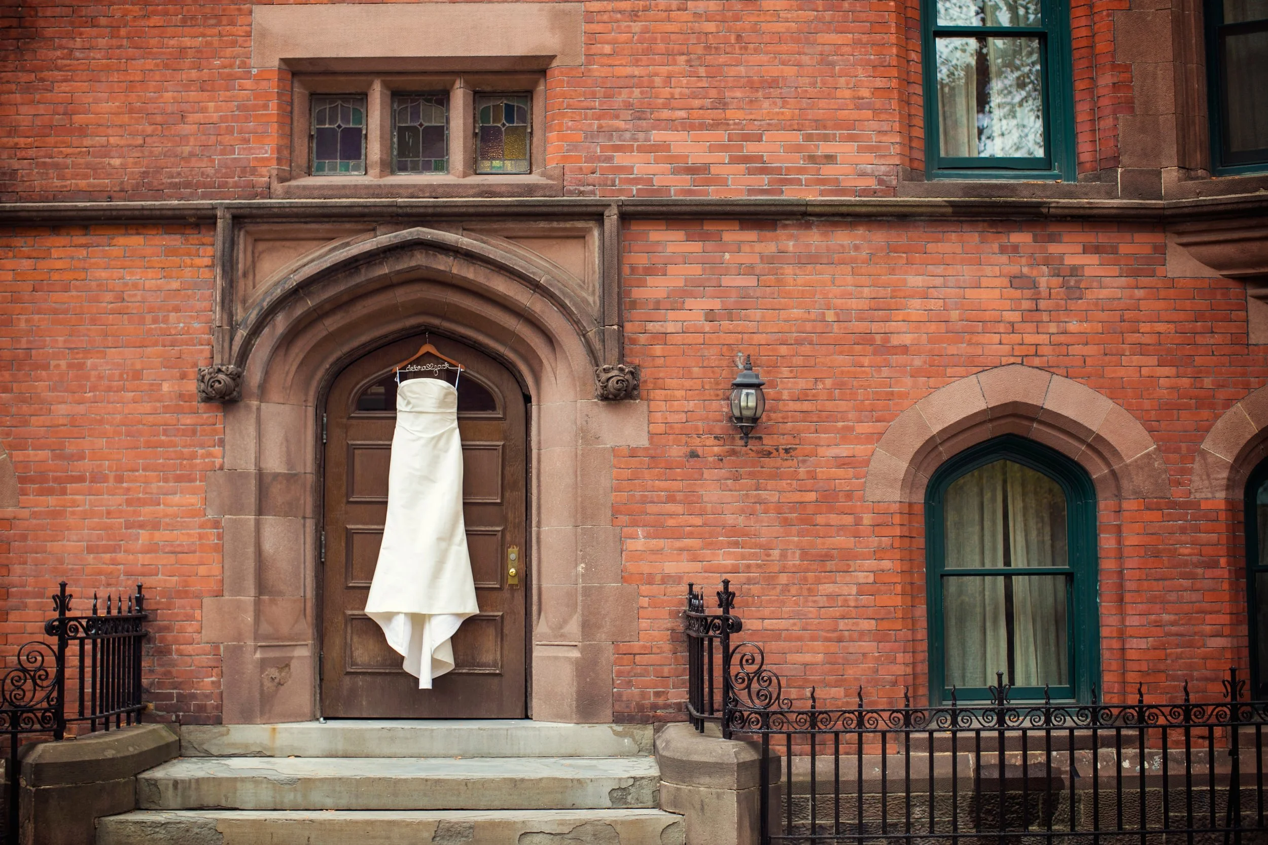 A white wedding dress hanging on the front door of a red brick building with arched windows and a stone staircase.