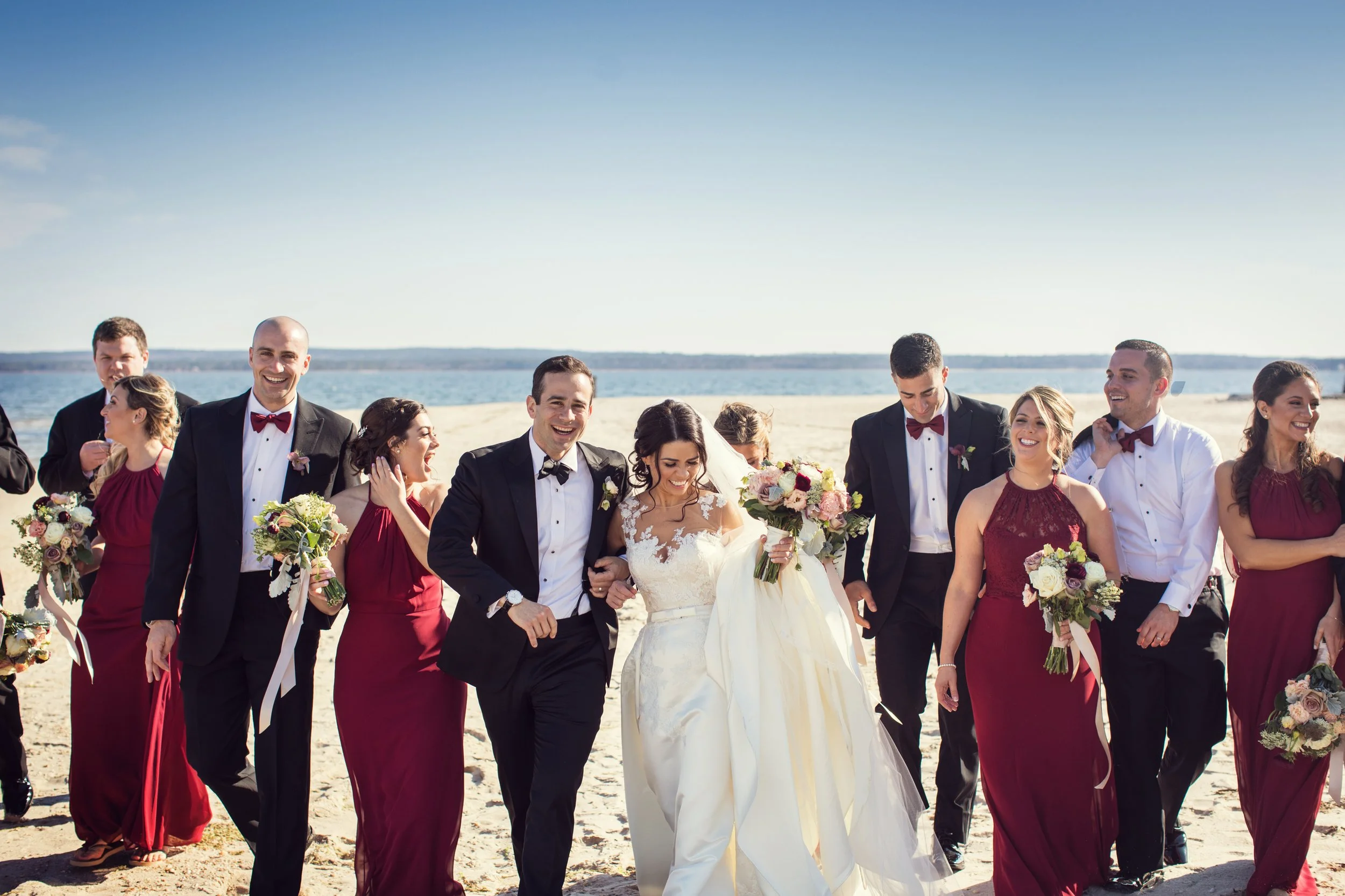 A group of wedding guests, including the bride and groom, walking on a beach with the ocean in the background. The bride is wearing a white wedding dress, and the groom is in a tuxedo. The bridesmaids are in burgundy dresses, and the groomsmen are in