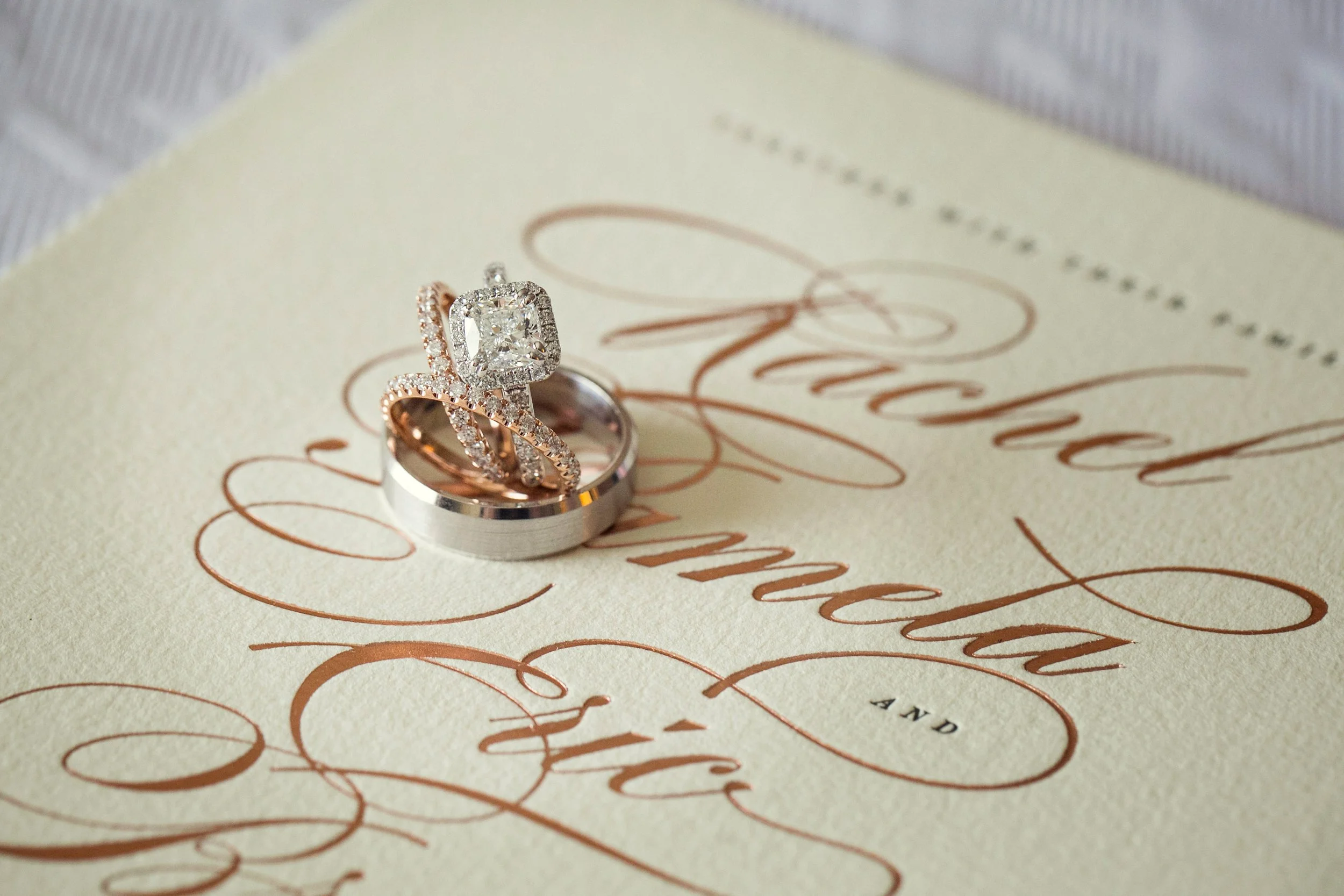 A wedding invitation with two rings on top, one with a large diamond and one with smaller diamonds, resting on the invitation with elegant cursive text.