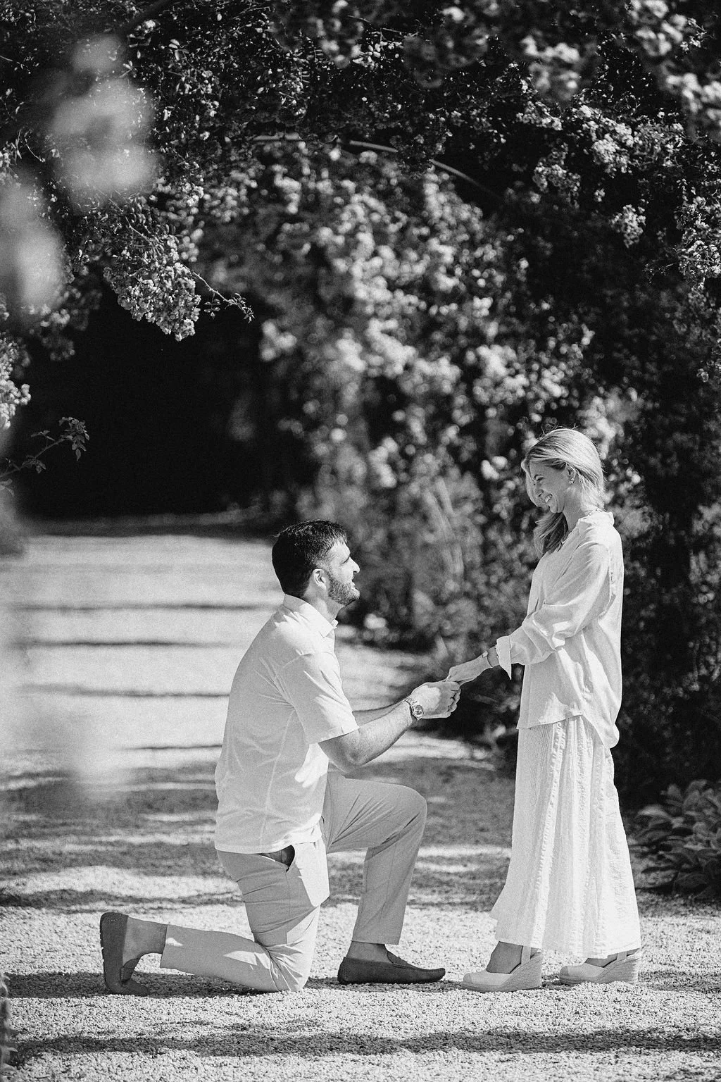 A man is kneeling on one knee, holding hands with a woman standing in front of him on a gravel pathway surrounded by trees and foliage, in a romantic outdoor setting.