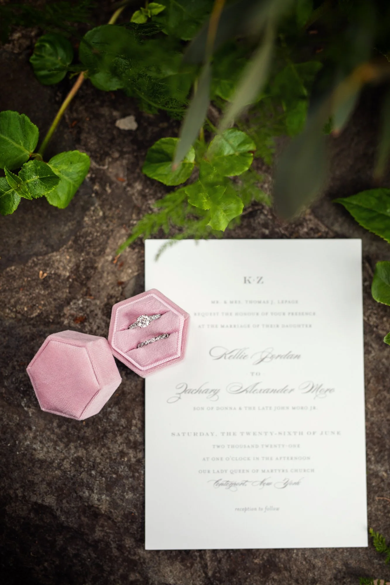Wedding invitation card with pink velvet ring box and two rings, surrounded by green foliage.